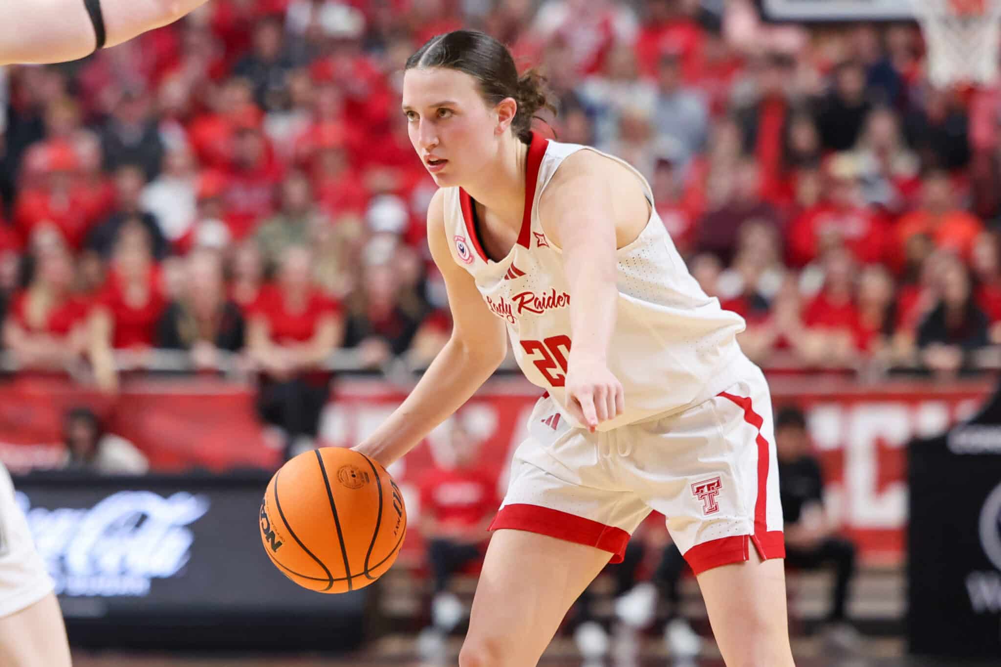 Female basketball player in a white-and-red uniform dribbles the ball during a game, focused on the play with a red-clad crowd in the background.
