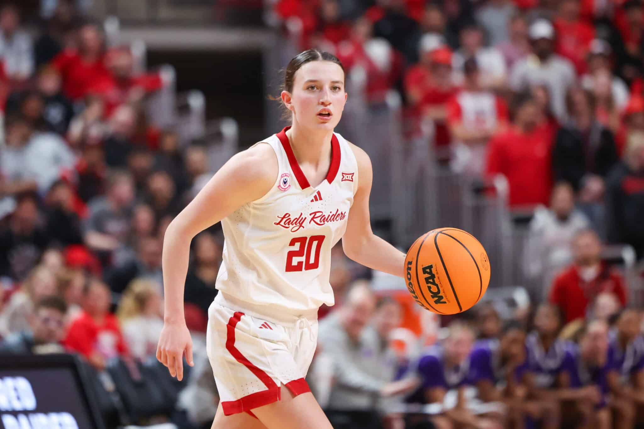 Lady Raiders guard wearing number 20 dribbles an orange basketball during a game with a crowded arena in the background.