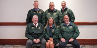 Six adults in matching green jackets pose for a group photo in a conference room.