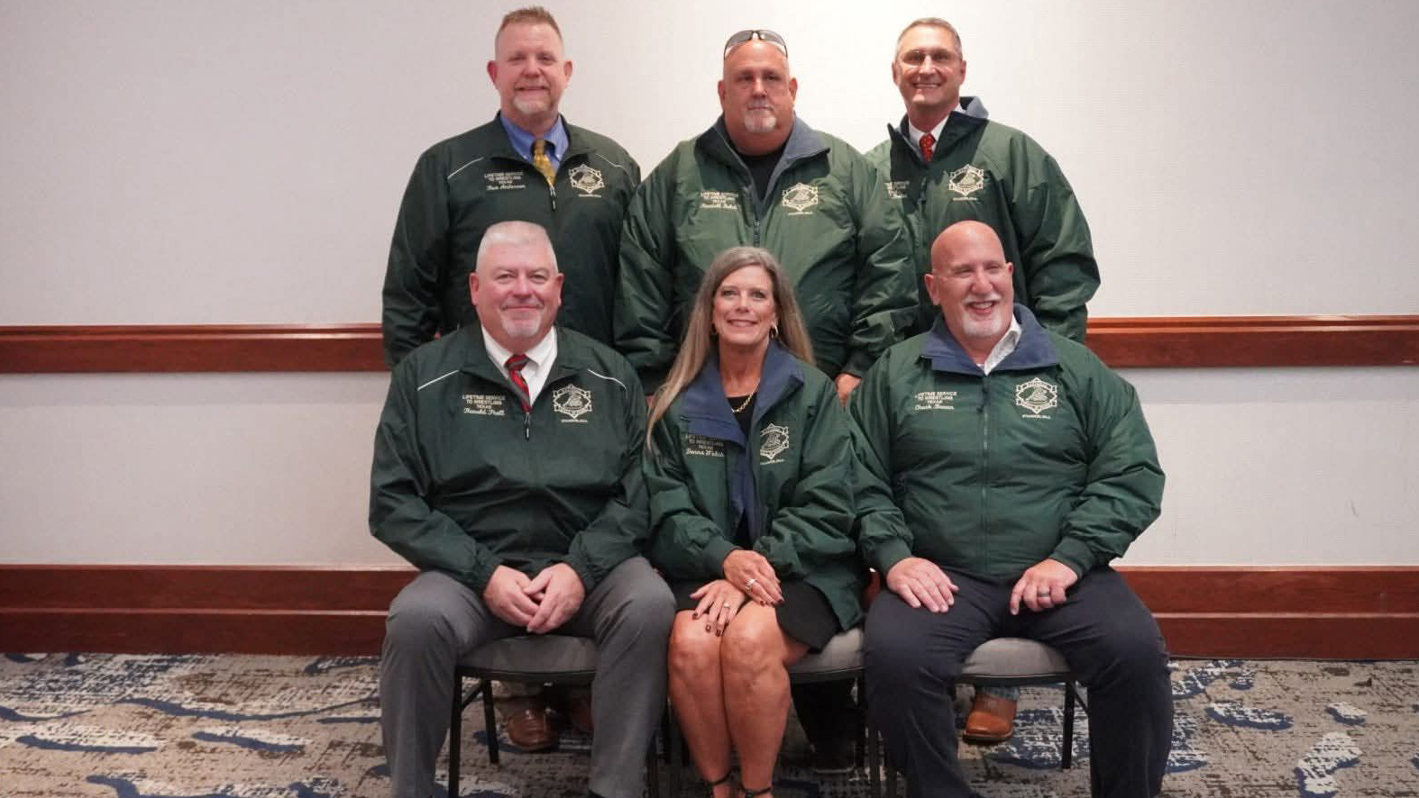 Six adults in matching green jackets pose for a group photo in a conference room.
