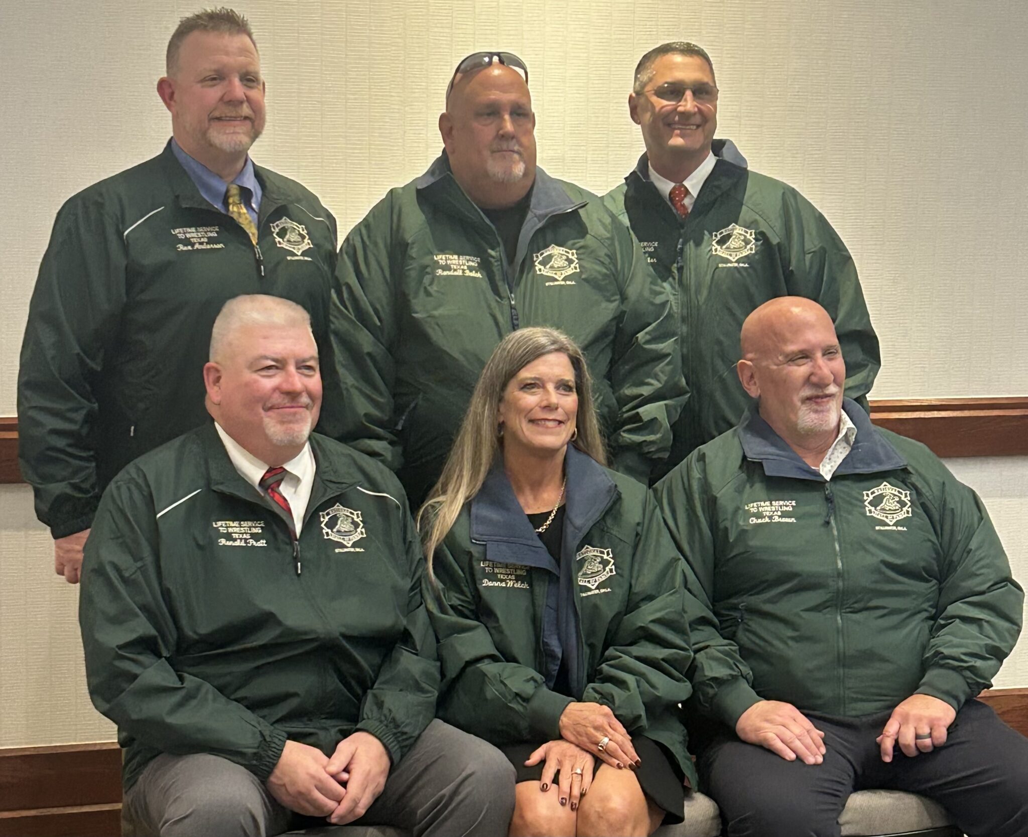 Six adults in matching green jackets posing for a group team photo in a neutral room with a bench backdrop.