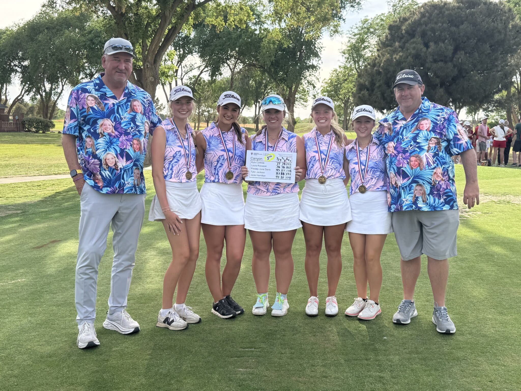 Seven golfers posing on a sunny course: two men in matching blue floral shirts flank five women in matching outfits with medals, one holding a scoreboard.