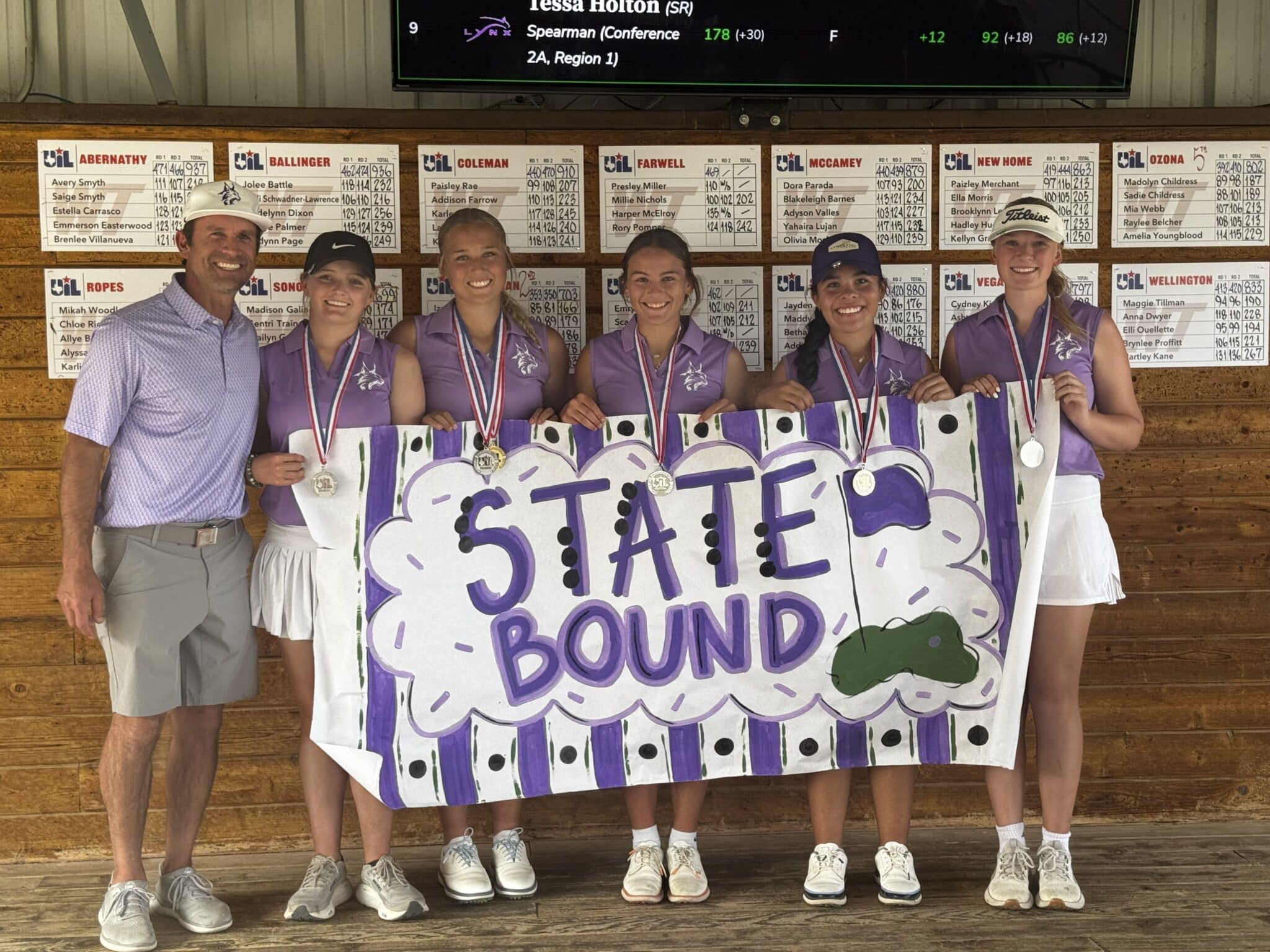 Six people pose indoors: a male coach and five female athletes in purple polo shirts and white skirts, wearing medals, holding a hand-painted banner reading 'STATE BOUND' with purple stripes, against a wooden wall with scoreboards behind them.