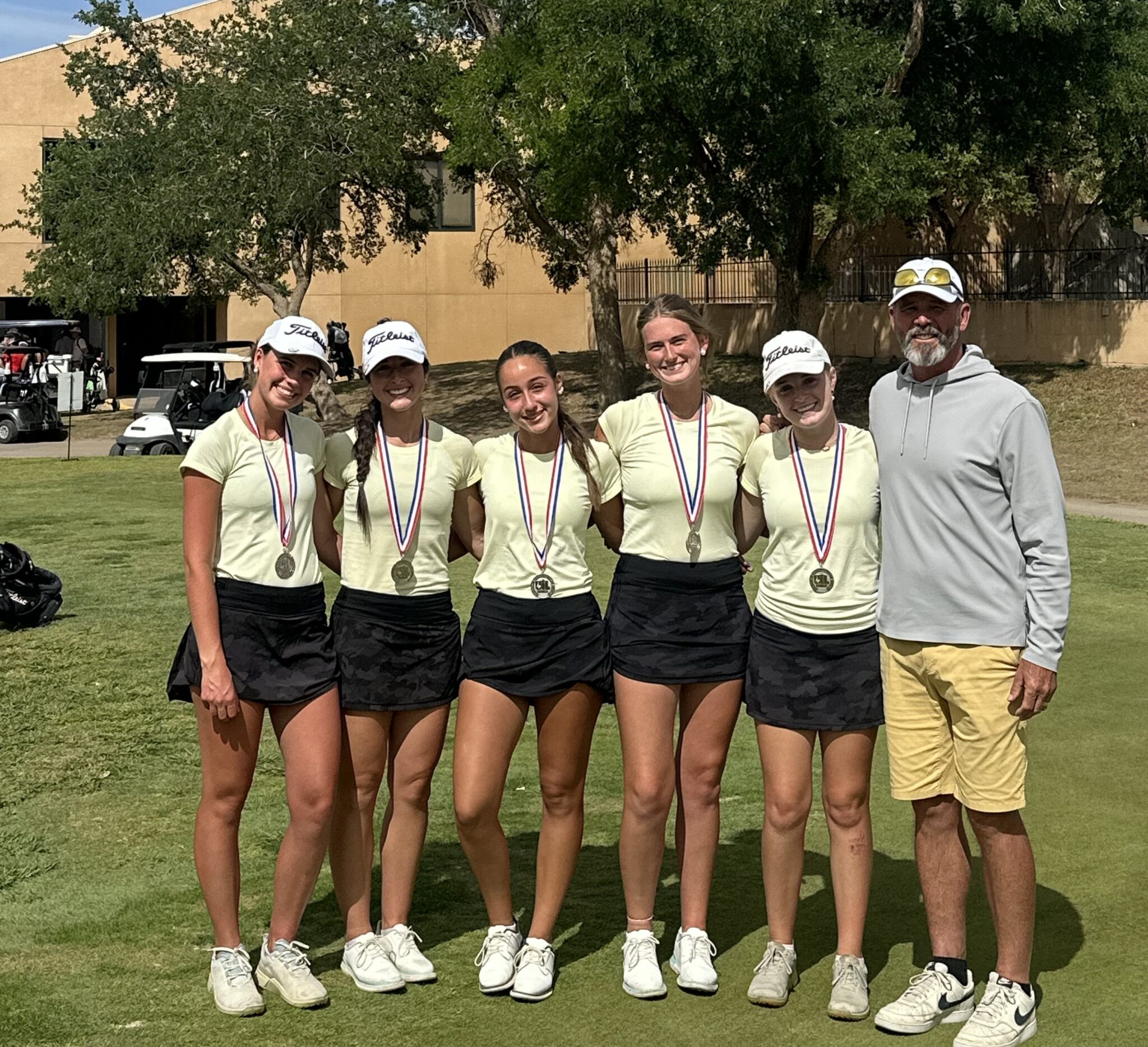 Five young golfers pose with medals on a green golf course, wearing light yellow shirts and black skirts, with a coach on the right.” ,