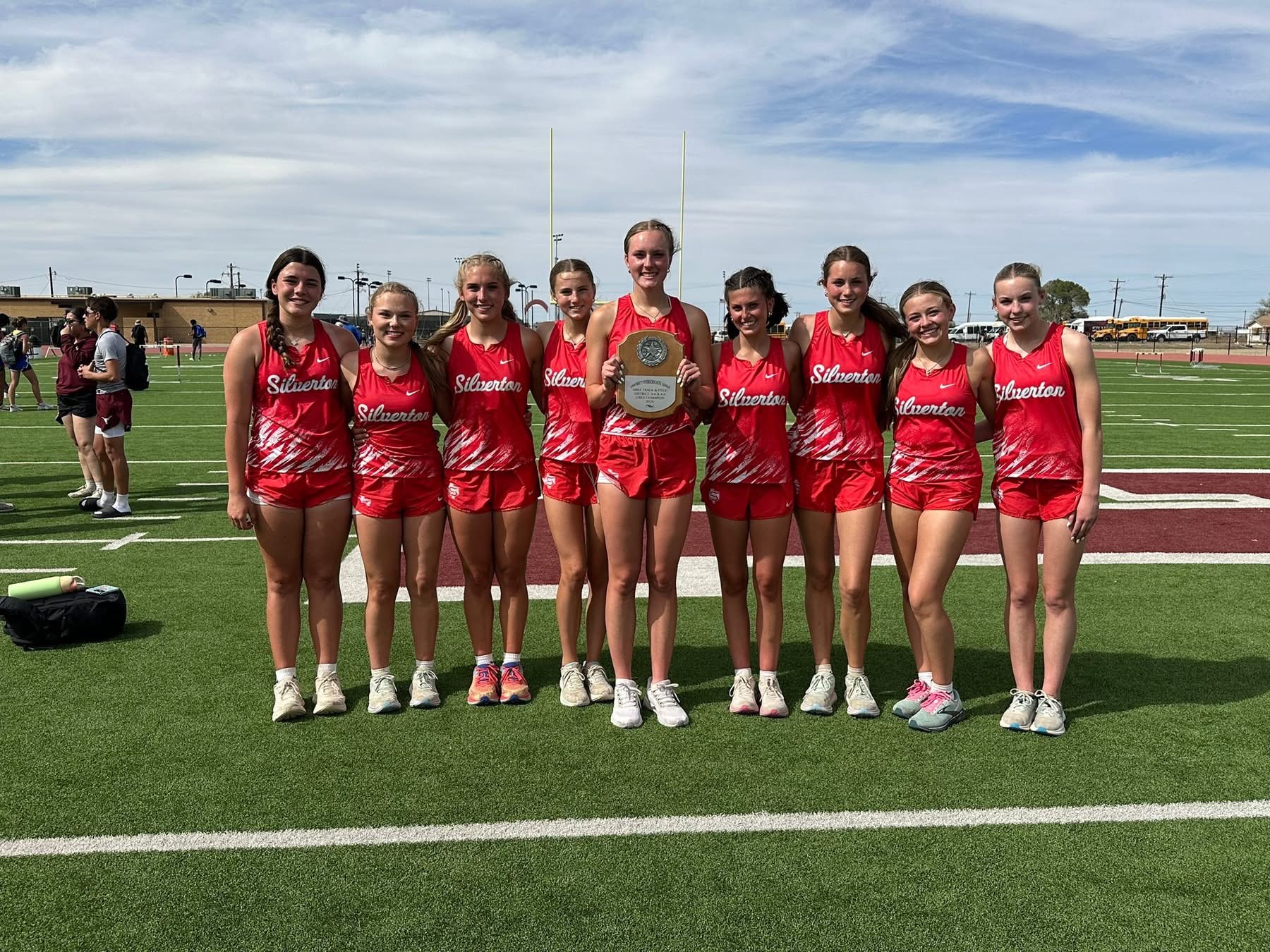 Girls track team in red uniforms posing with trophy on a football field after a meet. Silerton on jerseys.