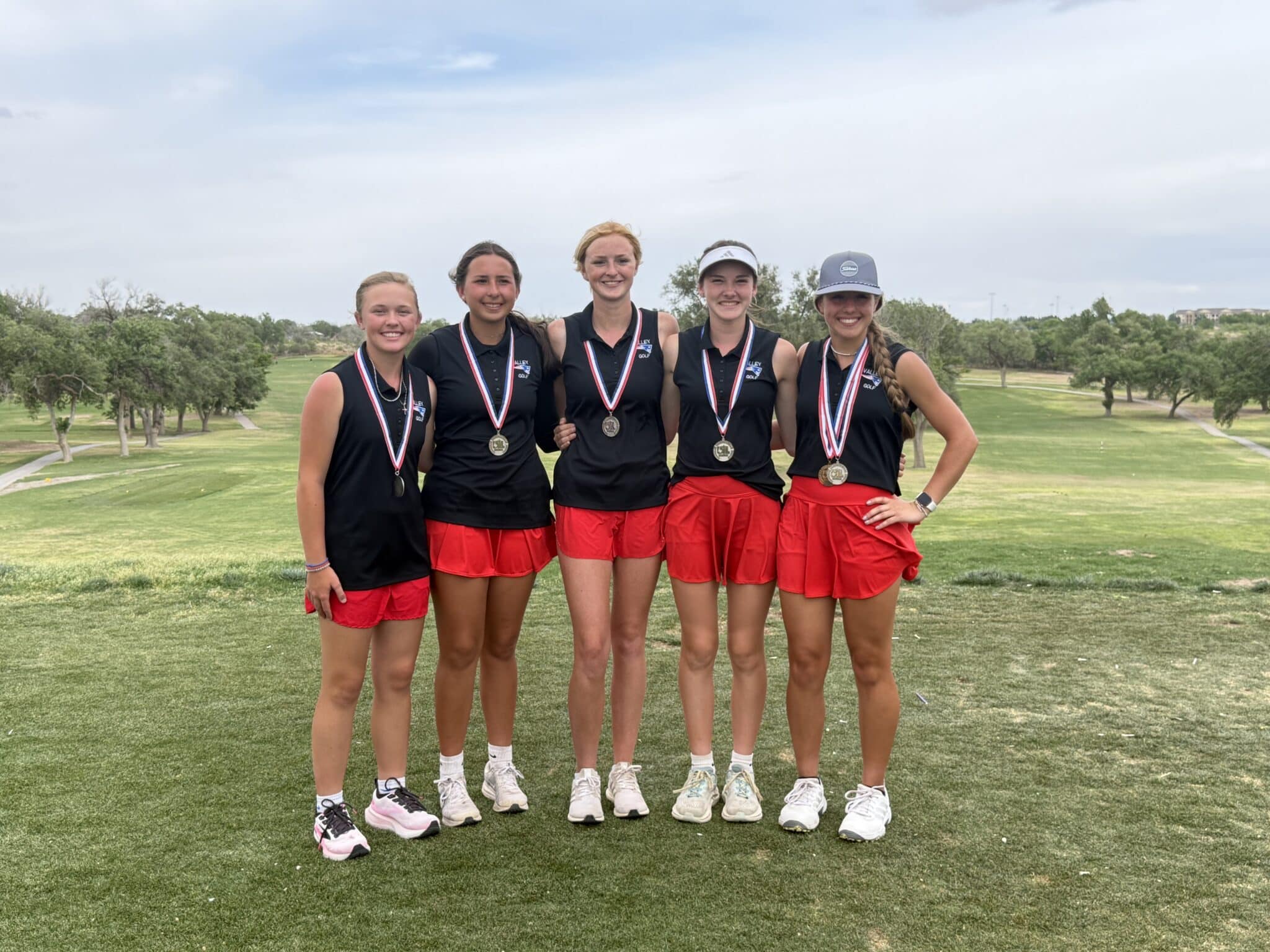 Five young female golfers stand together on a grassy golf course, wearing black tops, red shorts, and medals around their necks, smiling for a group photo.