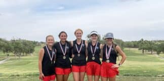 Five young female golfers stand together on a grassy golf course, wearing black tops, red shorts, and medals around their necks, smiling for a group photo.