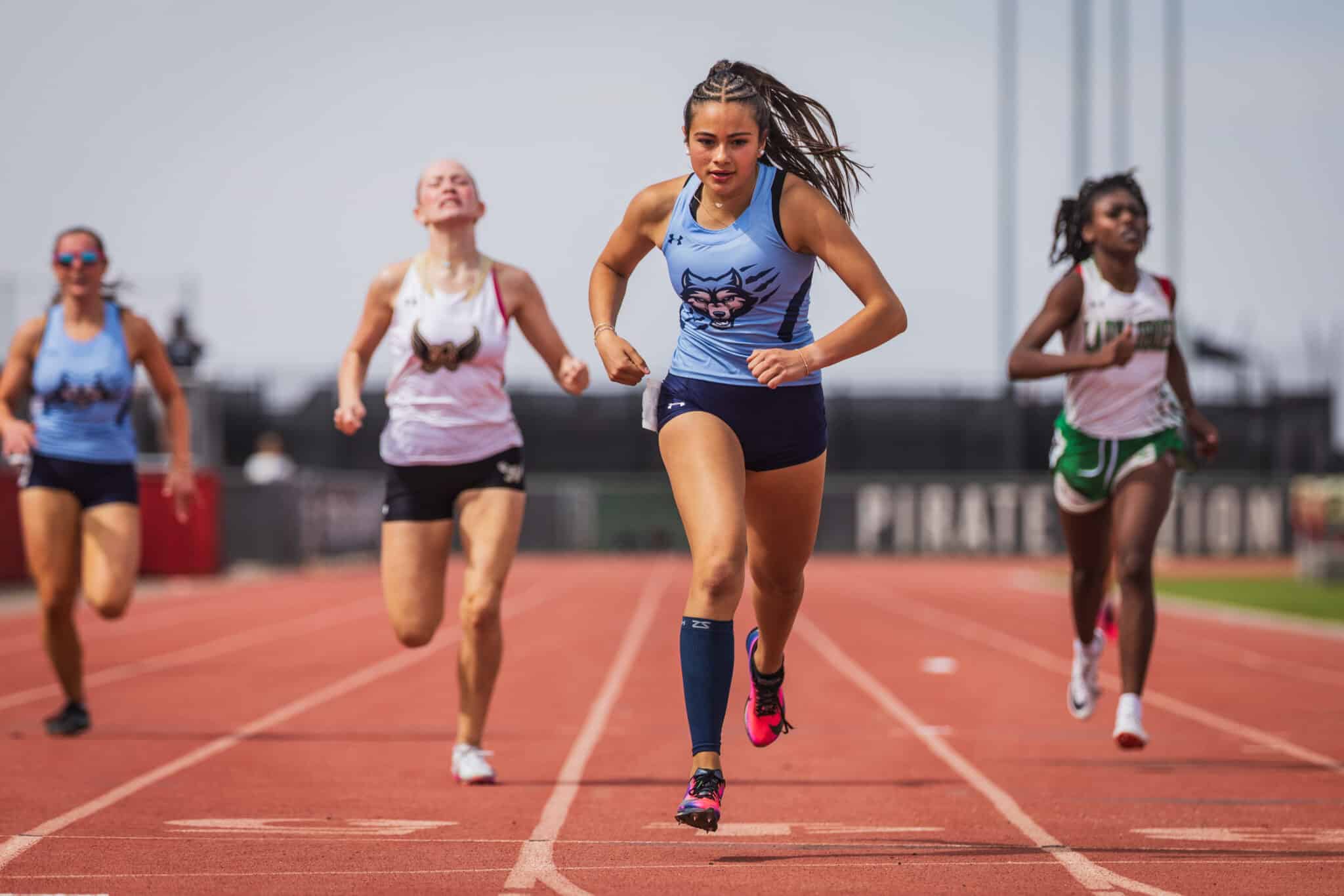 Young female sprinter in light blue top leading a race on a red track, others close behind on a sunny day.