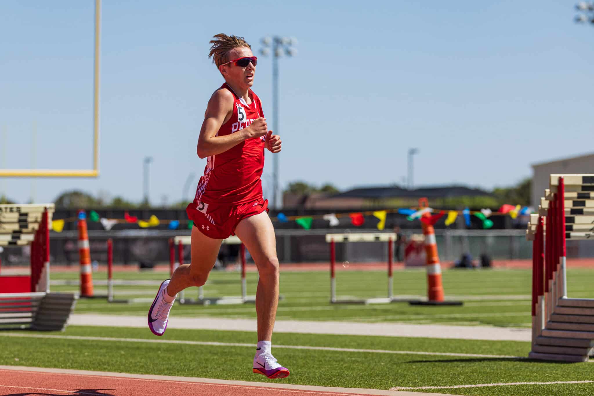 Young male sprinter in red uniform running on a track with hurdles in the background on a sunny day, wearing pink athletic shoes and red sunglasses.