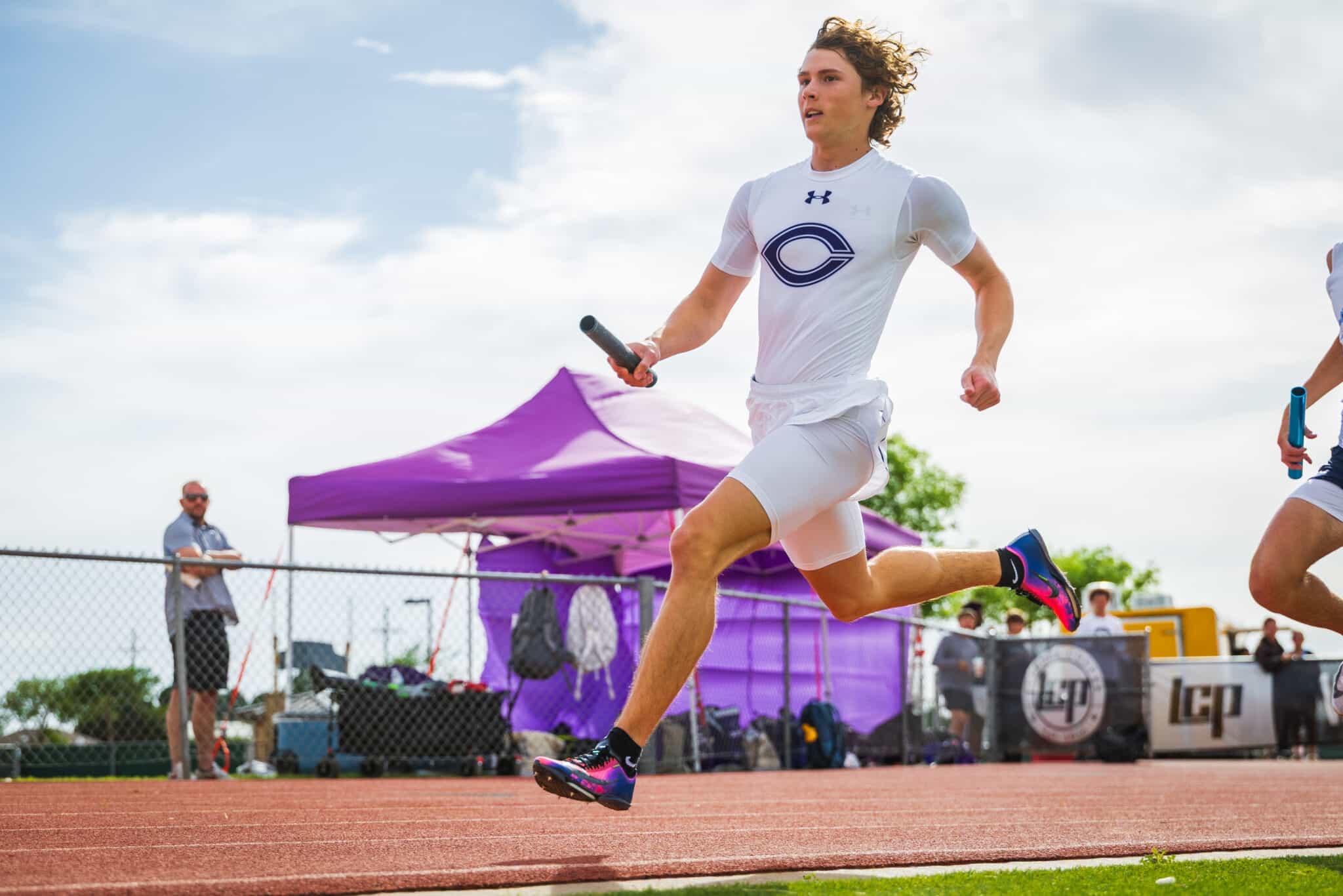 Male relay sprinter in a white uniform runs on a track, holding a baton, with a purple canopy and spectators in the background.