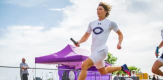 Male relay sprinter in a white uniform runs on a track, holding a baton, with a purple canopy and spectators in the background.