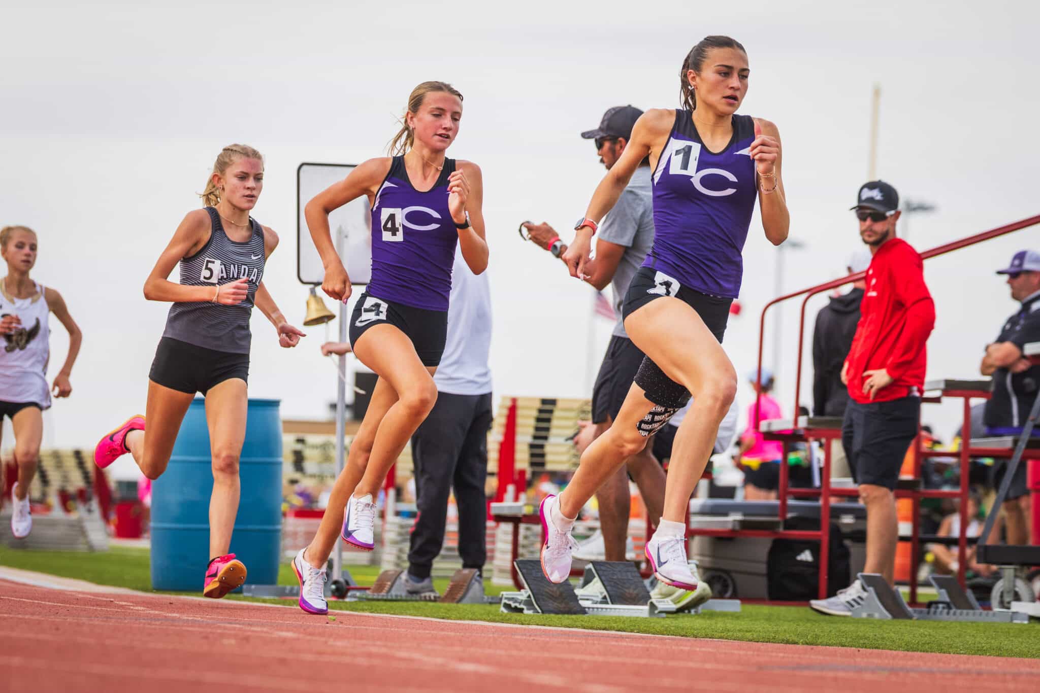 Group of young athletes sprinting on a red track, wearing numbered bibs and purple/gray uniforms, mid-race.