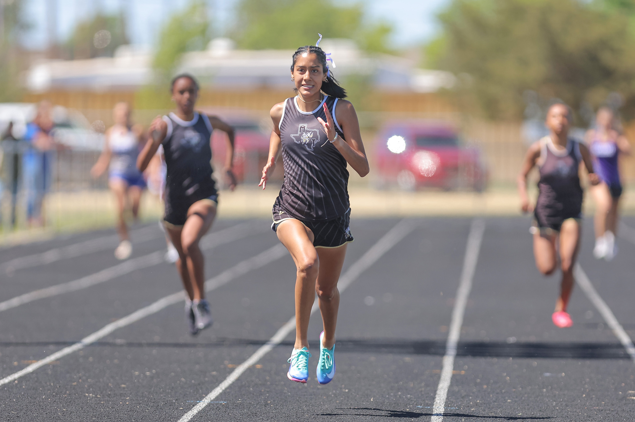 Female sprinter in a black uniform leads a track race on an outdoor field, turquoise shoes in mid-sprint with trailing runners behind.