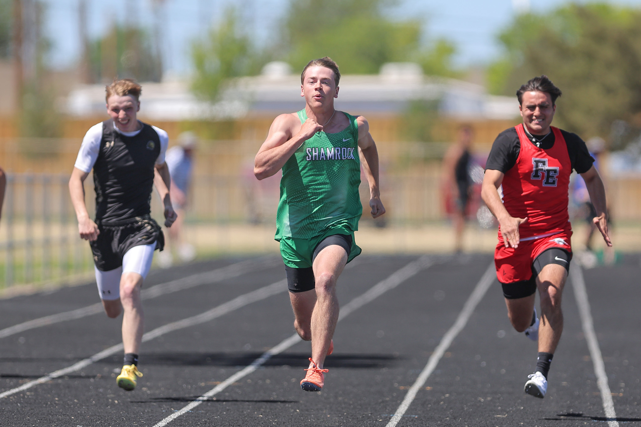 Three male sprinters race on an outdoor track, center in green Shamrock uniform leading, flanked by a black-and-white-clad runner left and a red-jersey runner right.