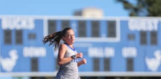 Female runner in a gray tank top and blue shorts sprinting on a track, with a blue school building in the background and a swinging ponytail.