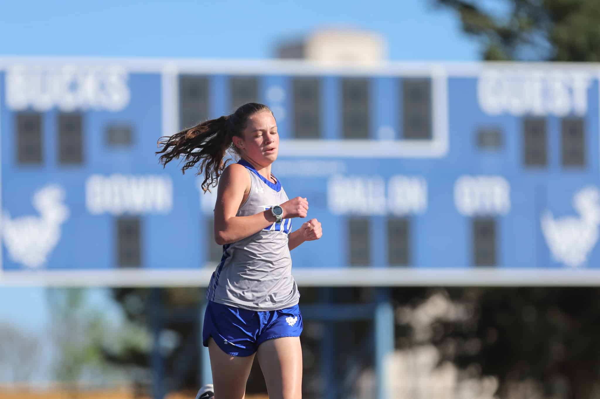 Female runner in a gray tank top and blue shorts sprinting on a track, with a blue school building in the background and a swinging ponytail.