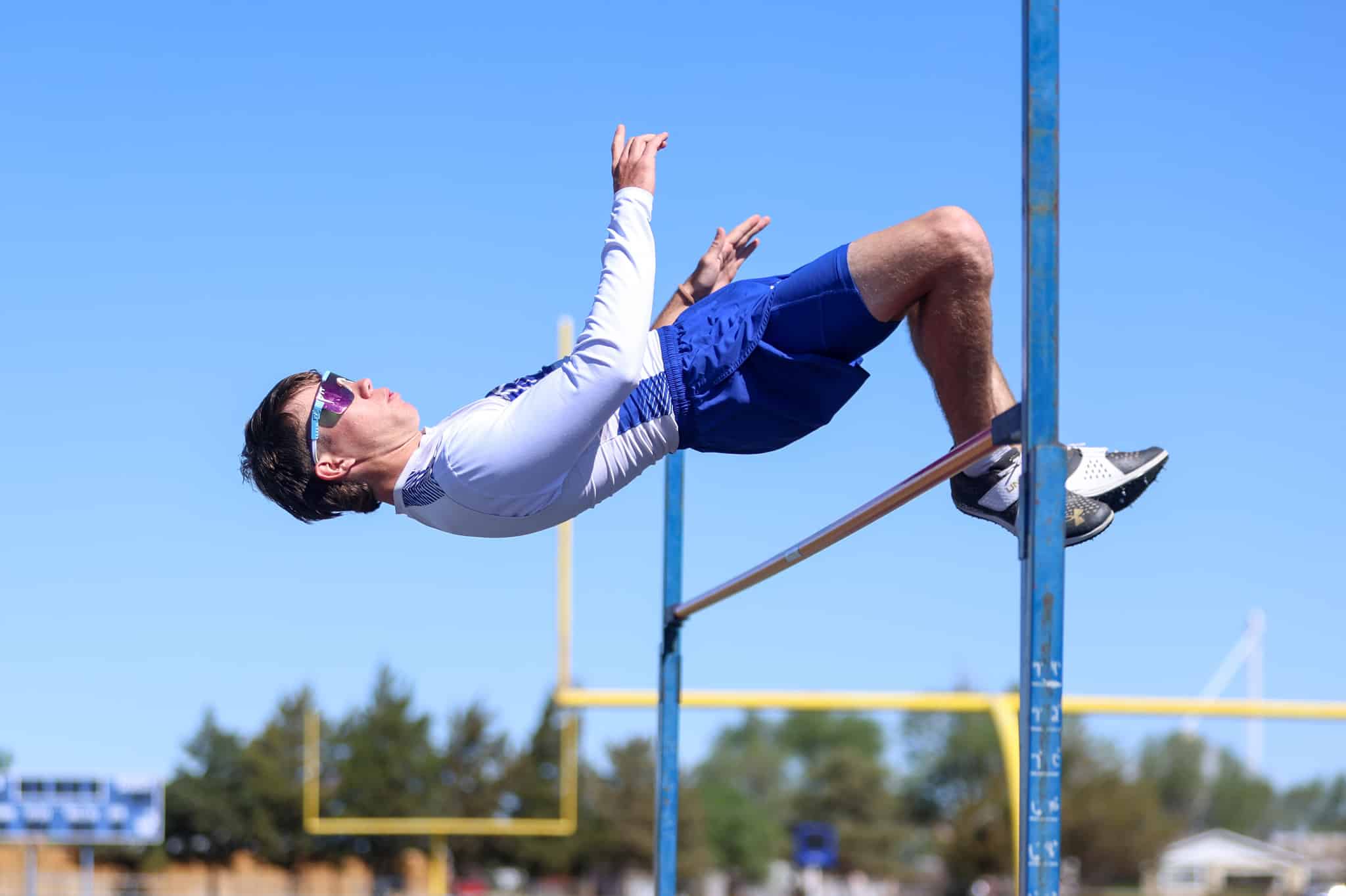 Male athlete mid-air during a high jump, arched back over the bar against a clear blue sky.