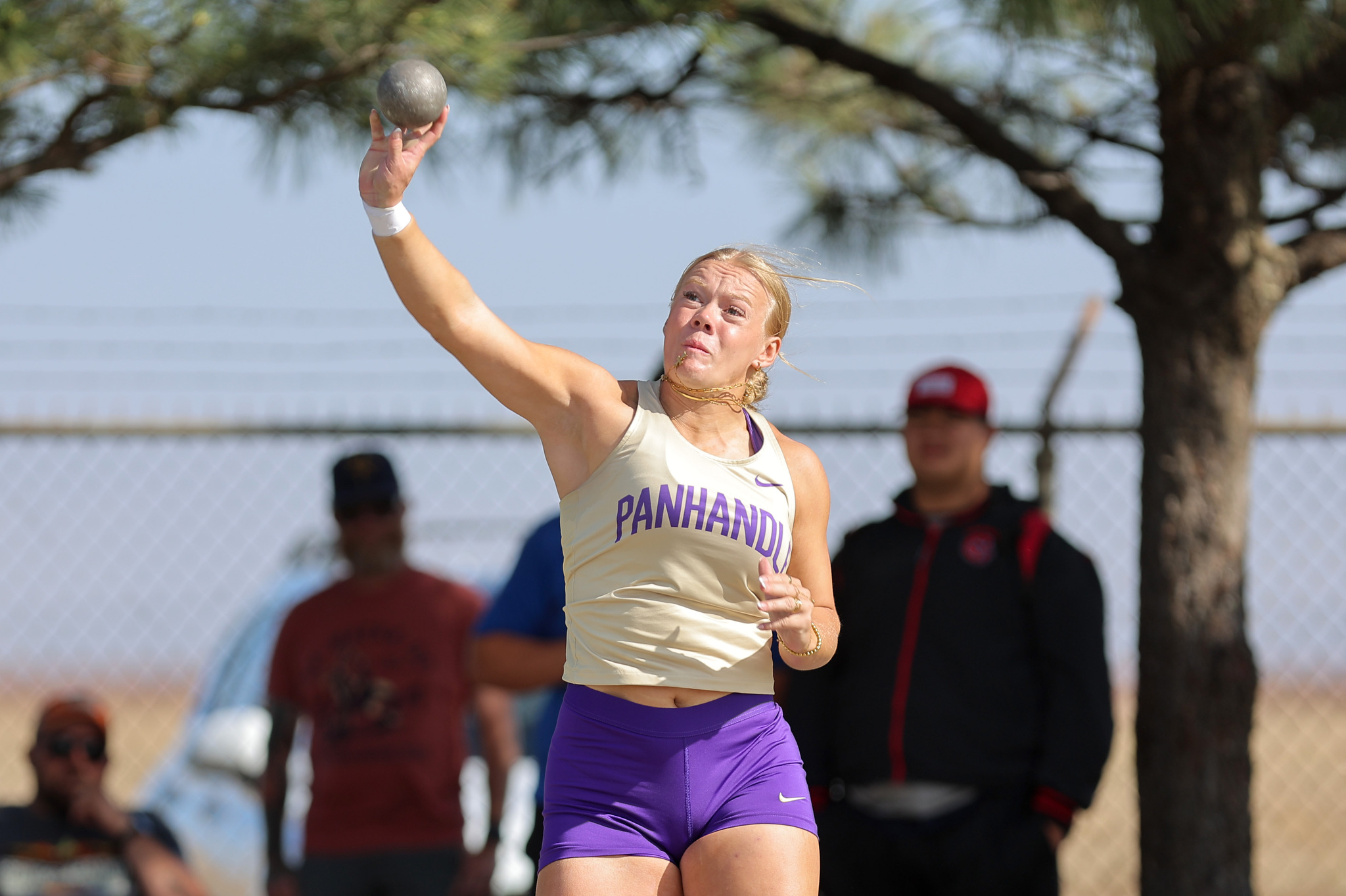 Athlete in a beige Panhandle top and purple shorts throwing a discus, with spectators and a chain-link fence in the background.