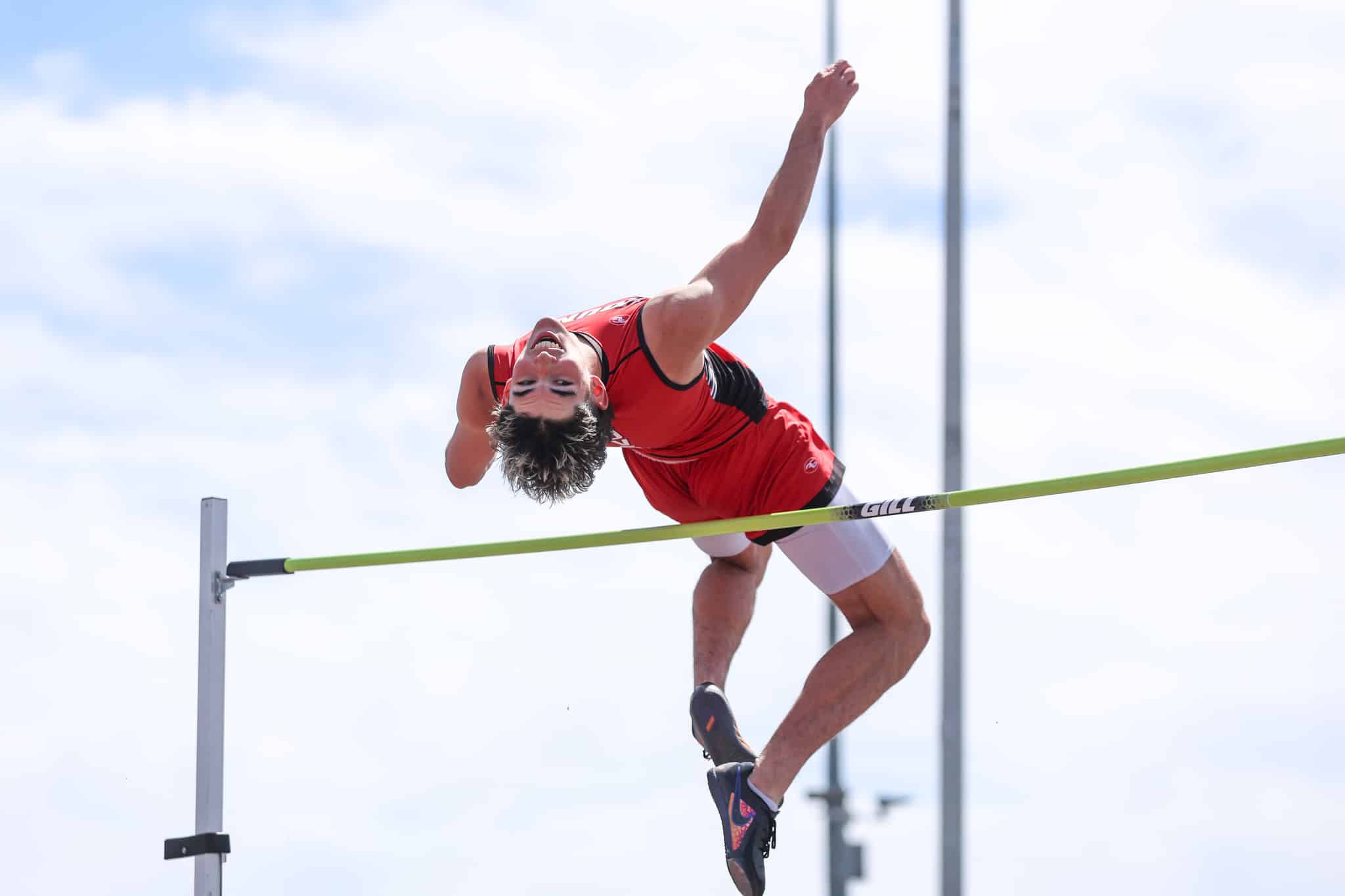 Male pole vaulter in a red uniform clears a bright green bar outdoors against a blue sky.