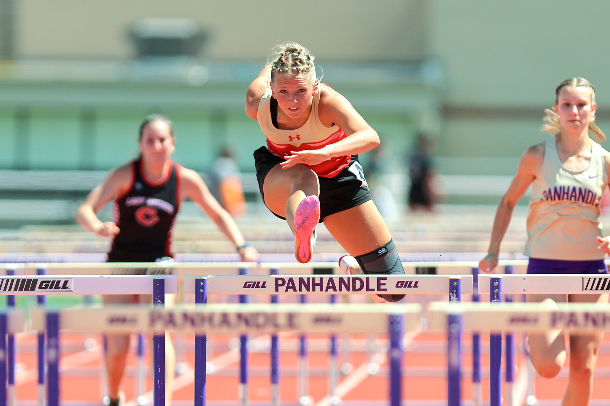 Female hurdler in orange and black mid-leap over a hurdle during a track race, with two competitors behind him/her in blur.