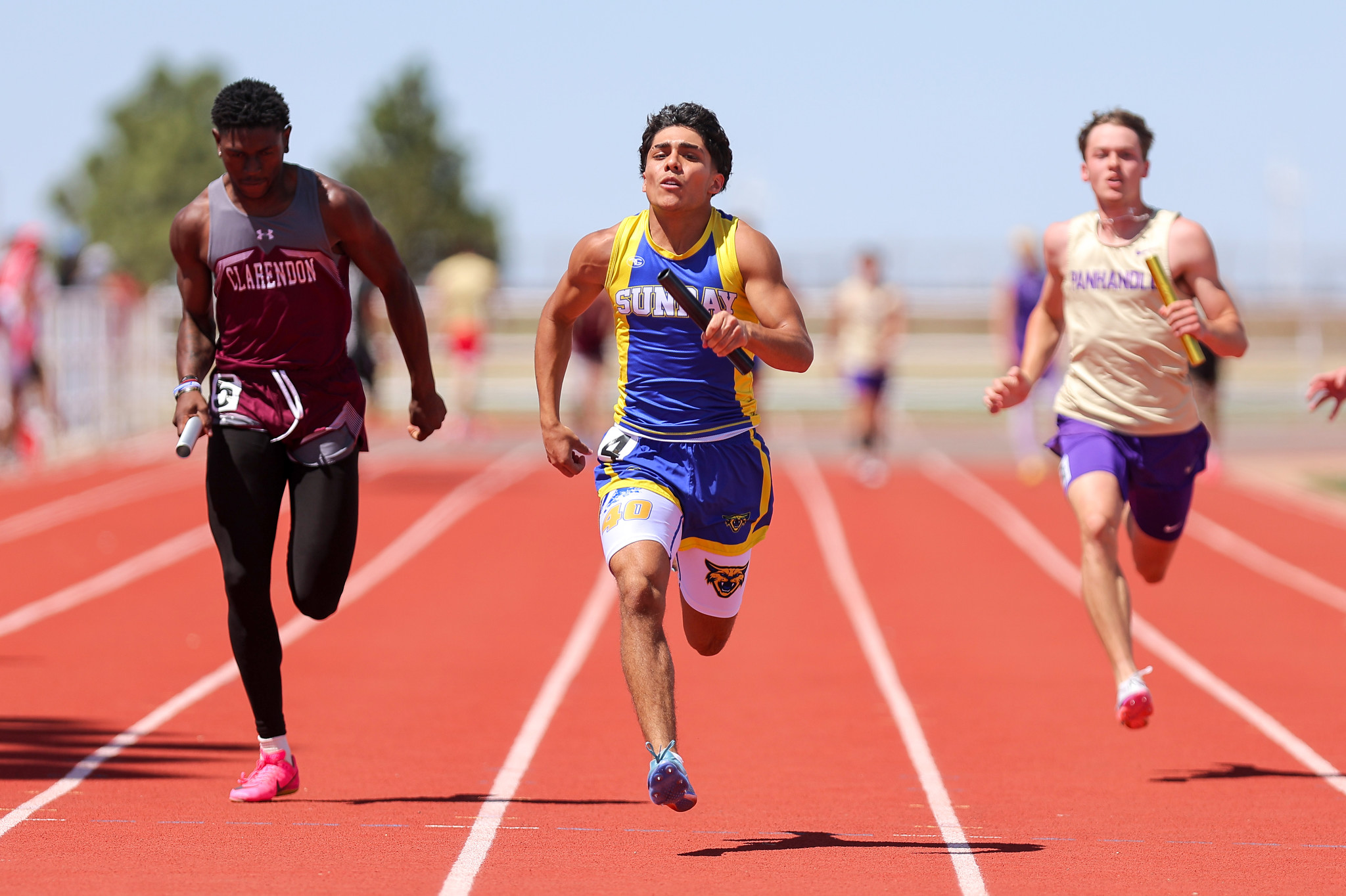 Three male sprinters race on a red track, leading with a blue‑and‑yellow uniform in the center as others trail behind.
