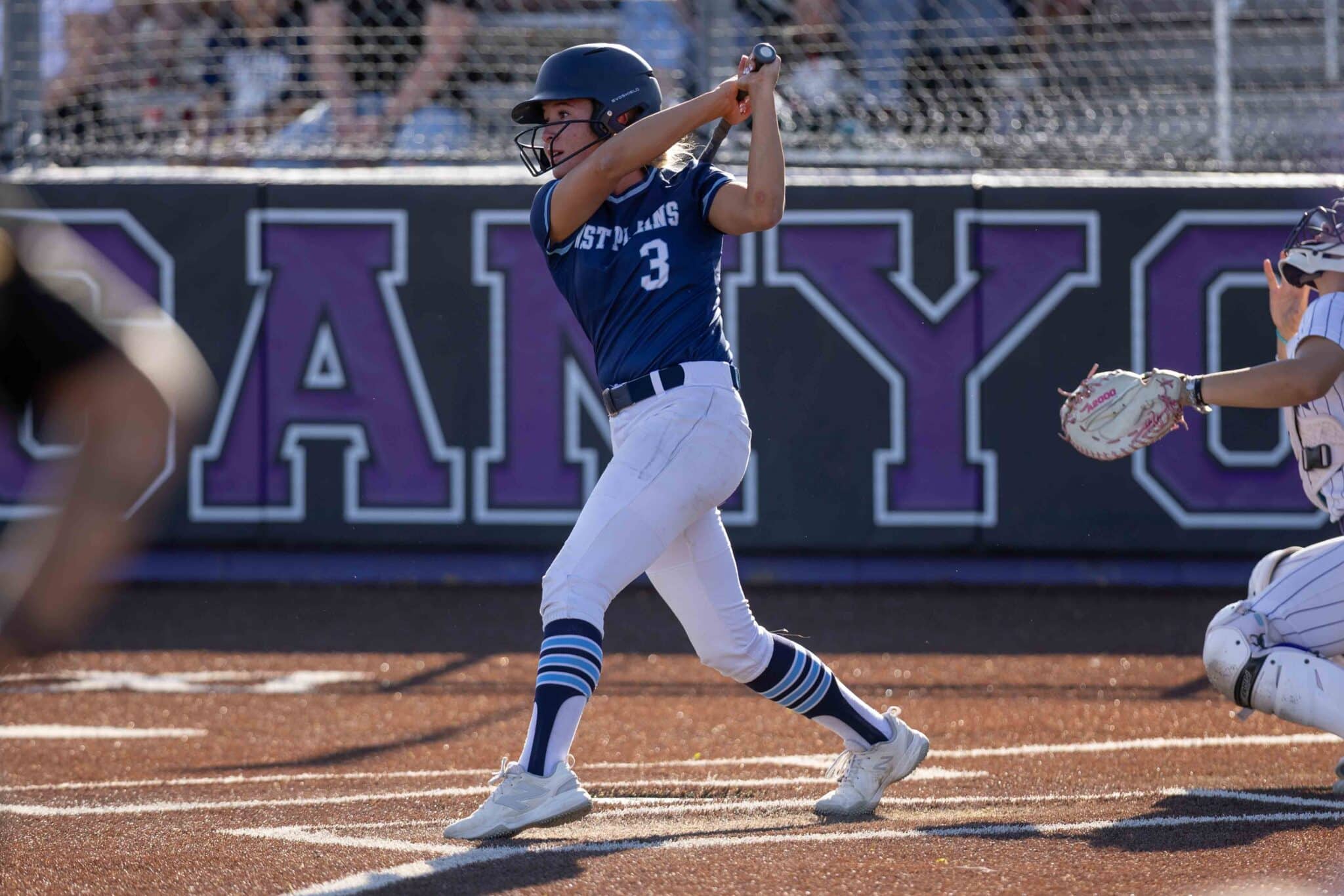 Female softball player in a blue uniform (number 3) swings at a pitch at home plate, catcher nearby behind the plate.