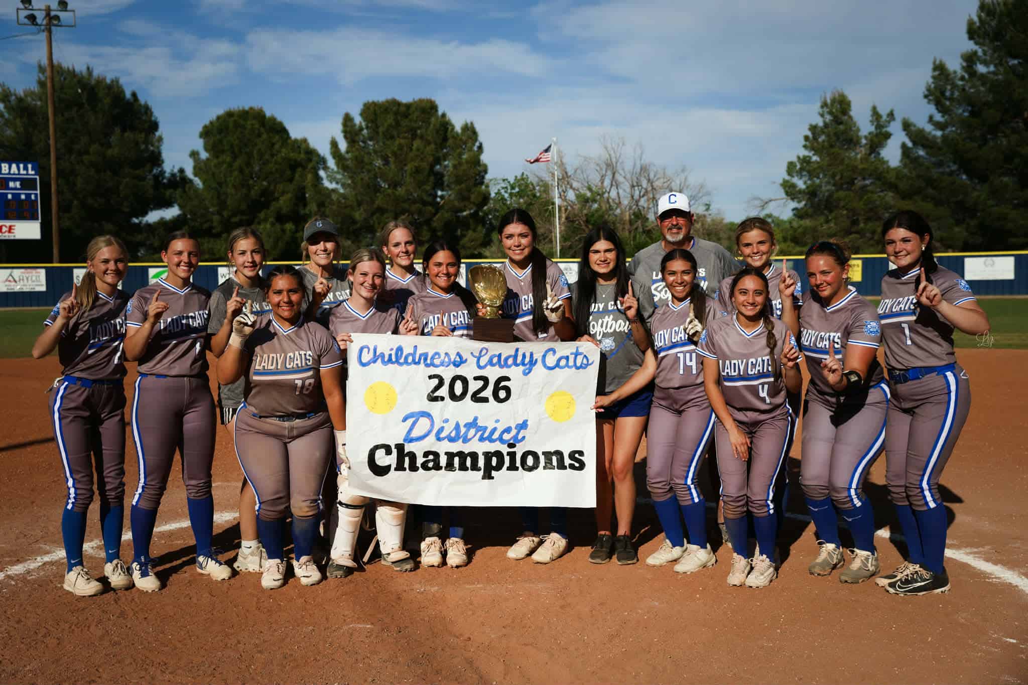 Girls softball team in gray uniforms posing with a trophy and a banner that says 'Childress Lady Cats 2026 District Champions' on a baseball field.