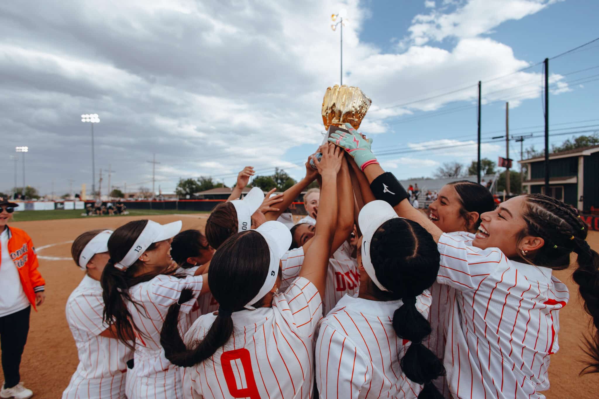 Girls softball players in white pinstriped uniforms lift a gold trophy in a celebratory huddle on the dirt field. Looks like a championship moment.