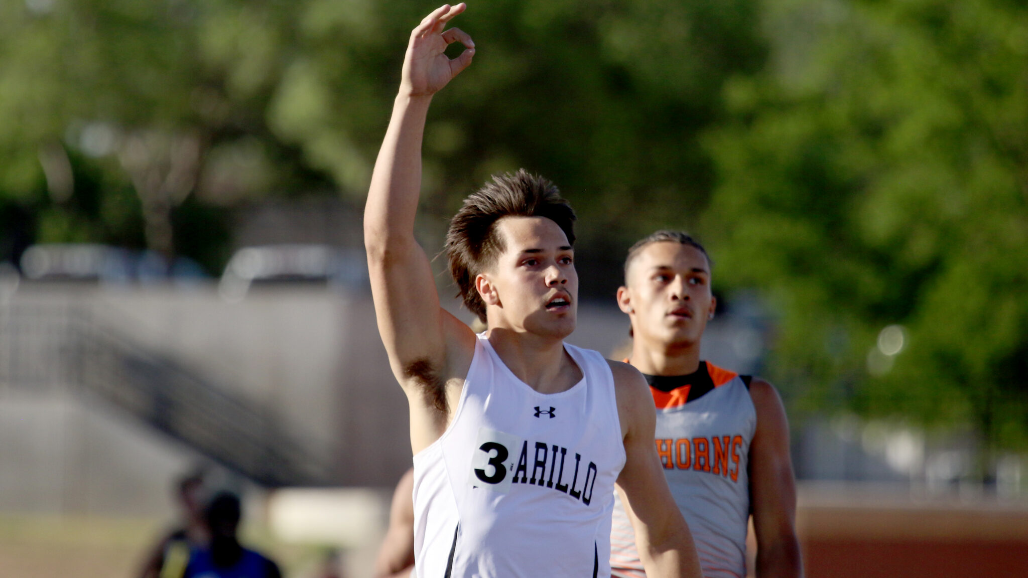 Sprinter in a white jersey raises his right arm in celebration as he leads a track race, with a rival in gray behind him.