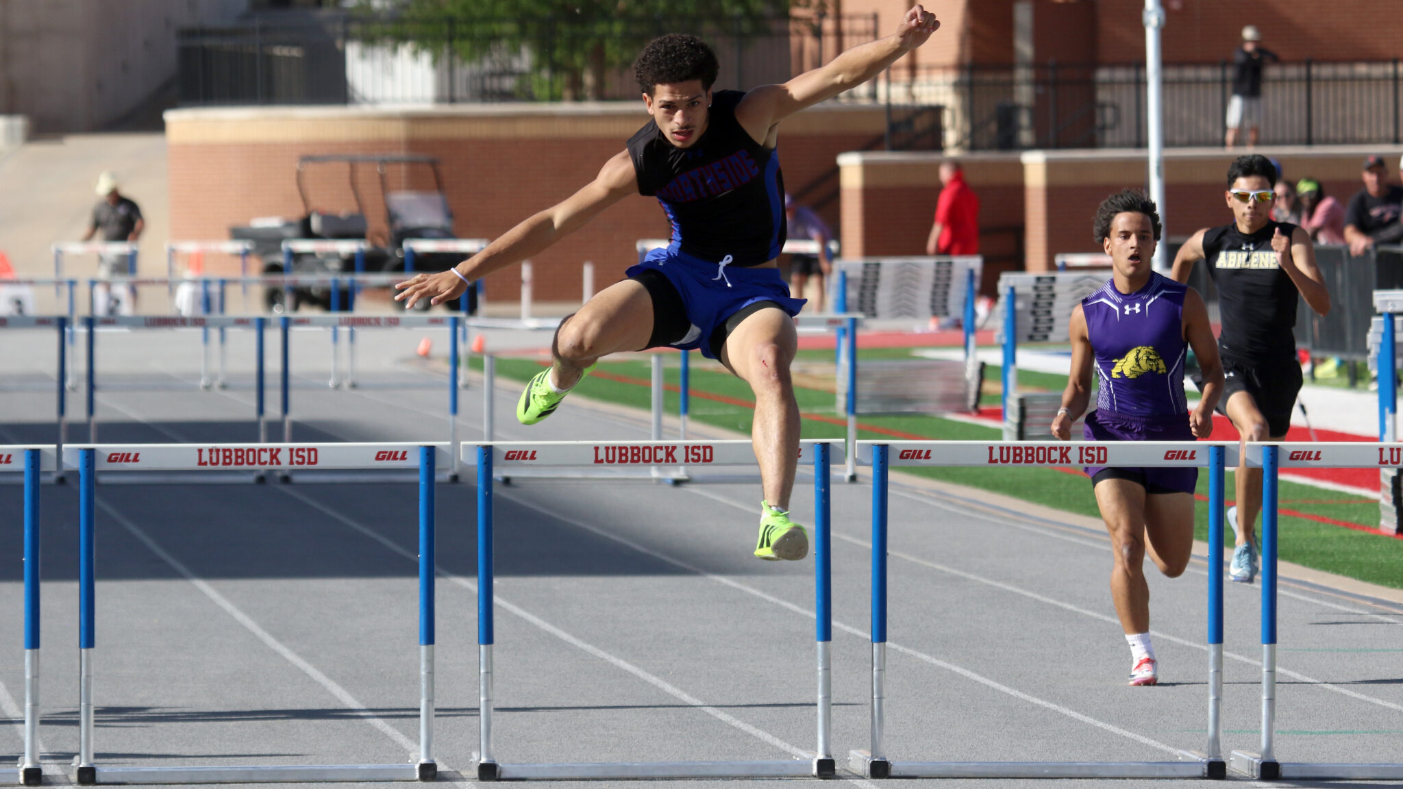 Athlete clears a hurdle mid-air on a track, wearing a black singlet and blue shorts; neon green shoes stand out.