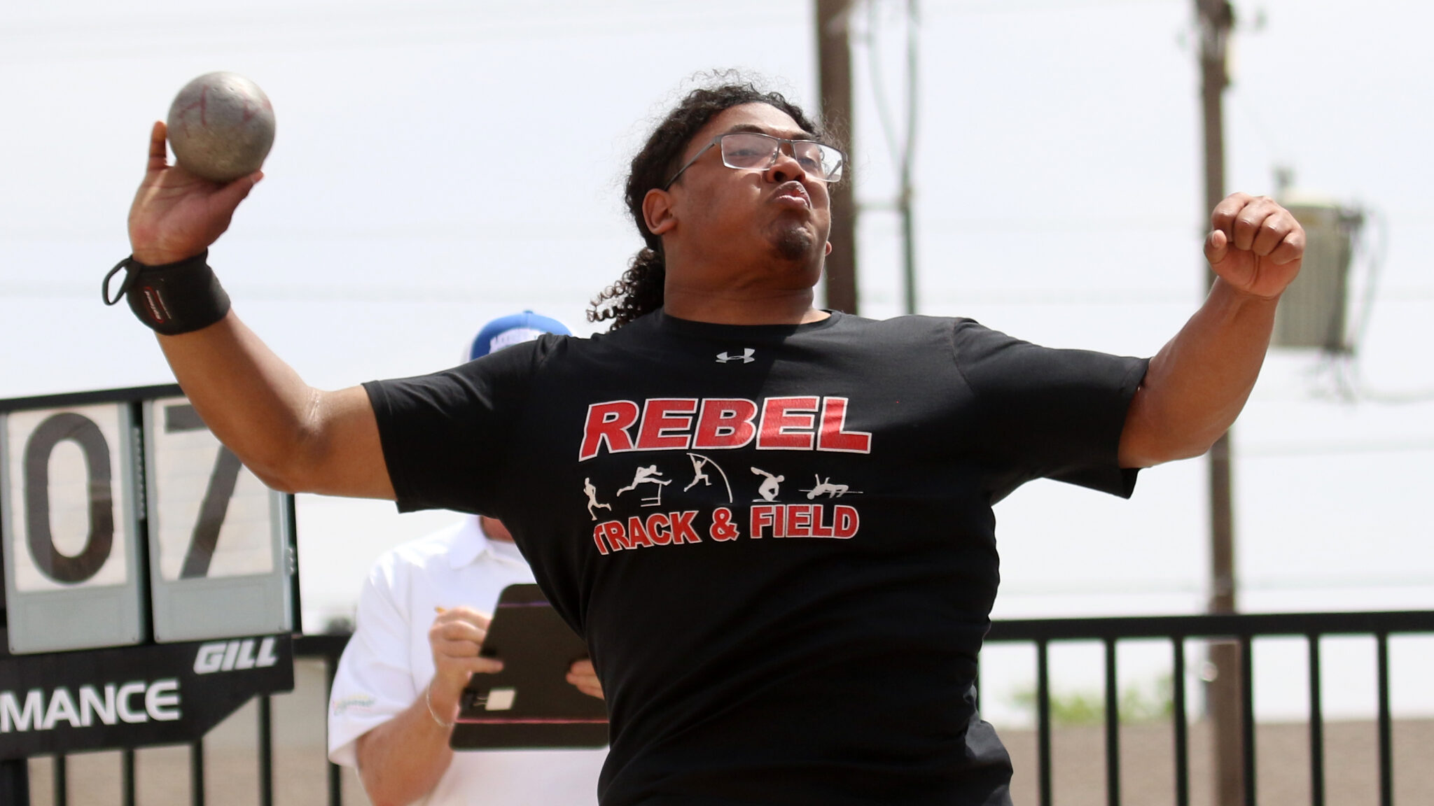 Male athlete in a black 'REBEL TRACK & FIELD' shirt throwing a shot put outdoors, focused on the throw.