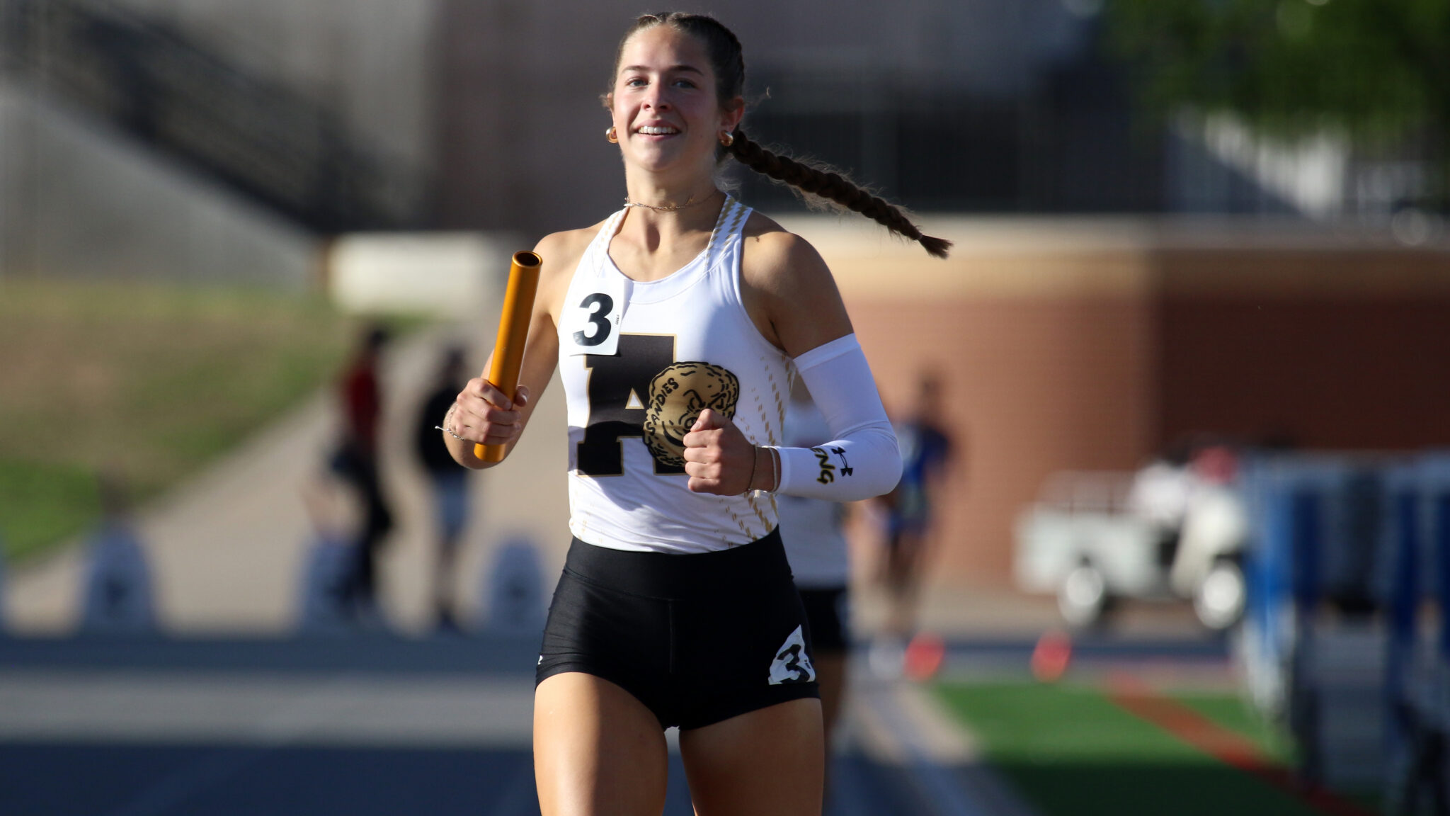 Female relay runner sprinting on a track, holding an orange baton and wearing a white top with a black number 3 bib.