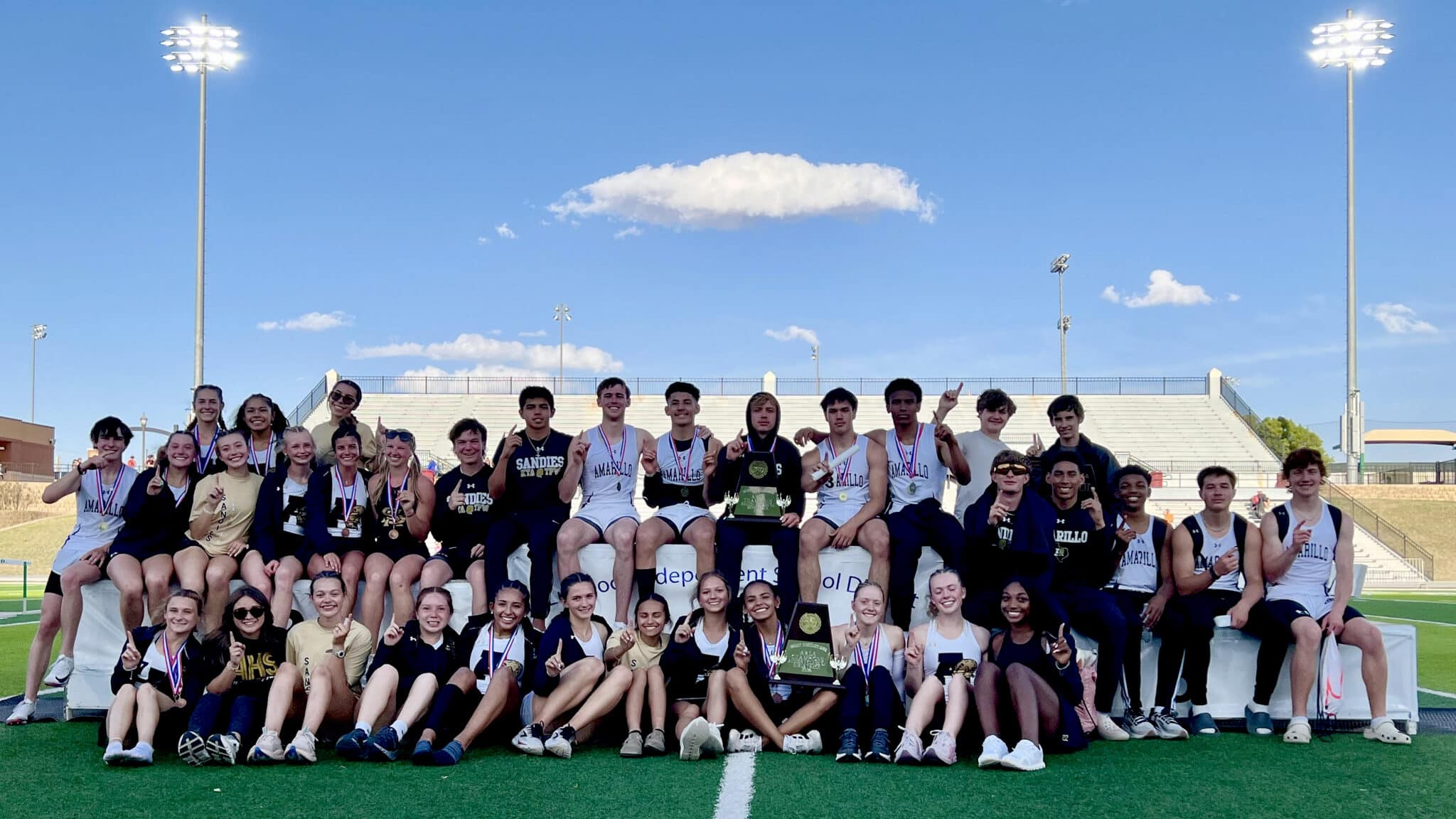 Large group of young athletes on a podium at a track stadium, wearing medals and posing for a team photo.