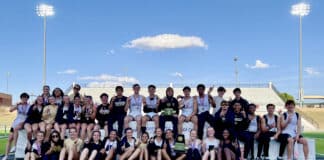 Large group of young athletes on a podium at a track stadium, wearing medals and posing for a team photo.