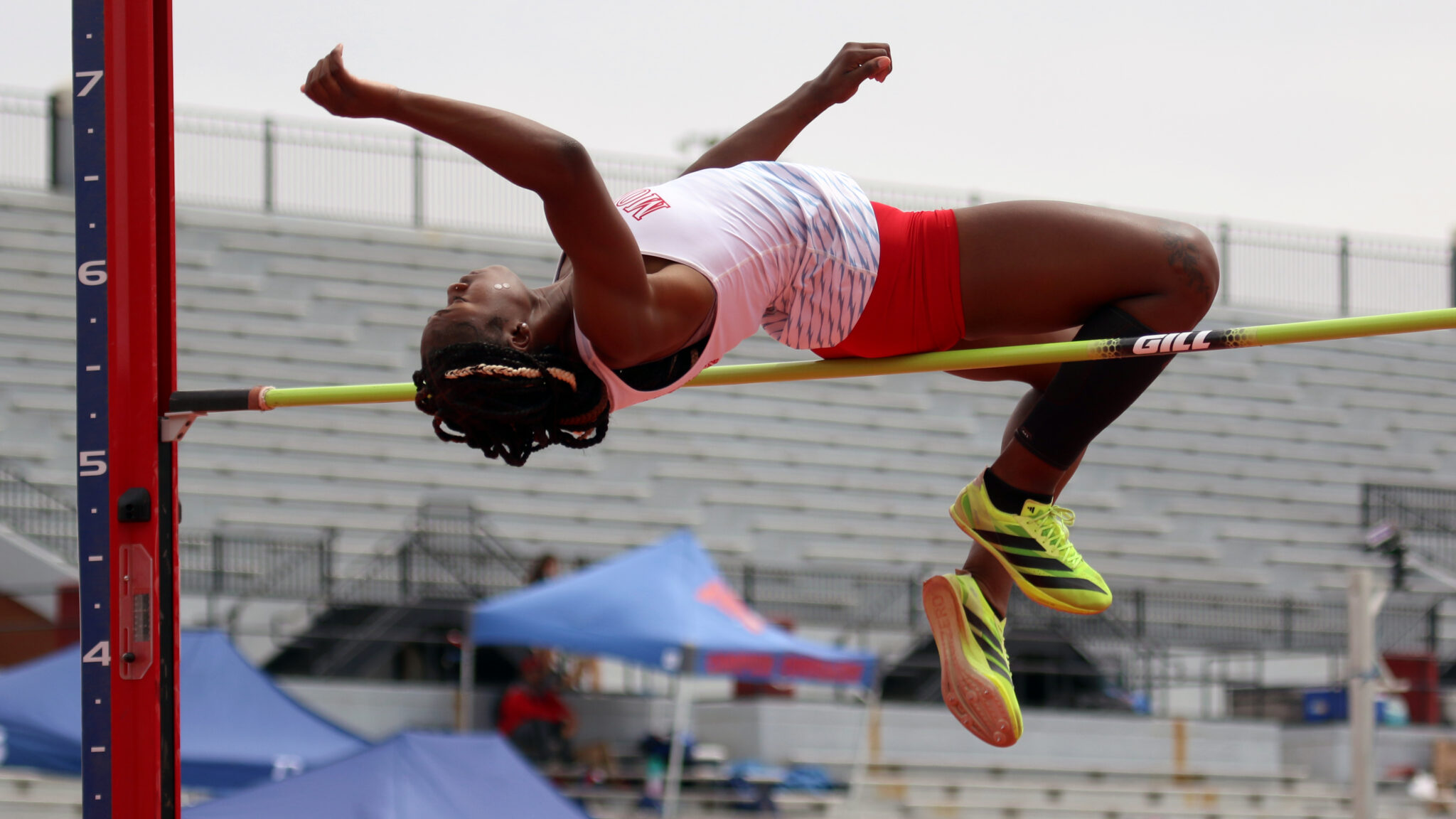 Female pole vaulter in white and red uniform clears a bar mid-air at a stadium.