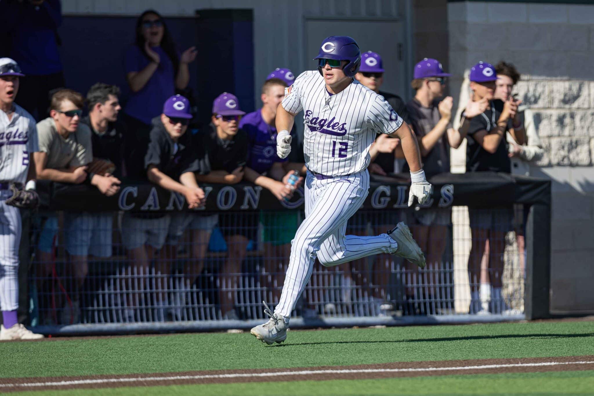 Baseball player in white pinstripe uniform (#12) sprints along the basepath past cheering teammates in purple helmets.
