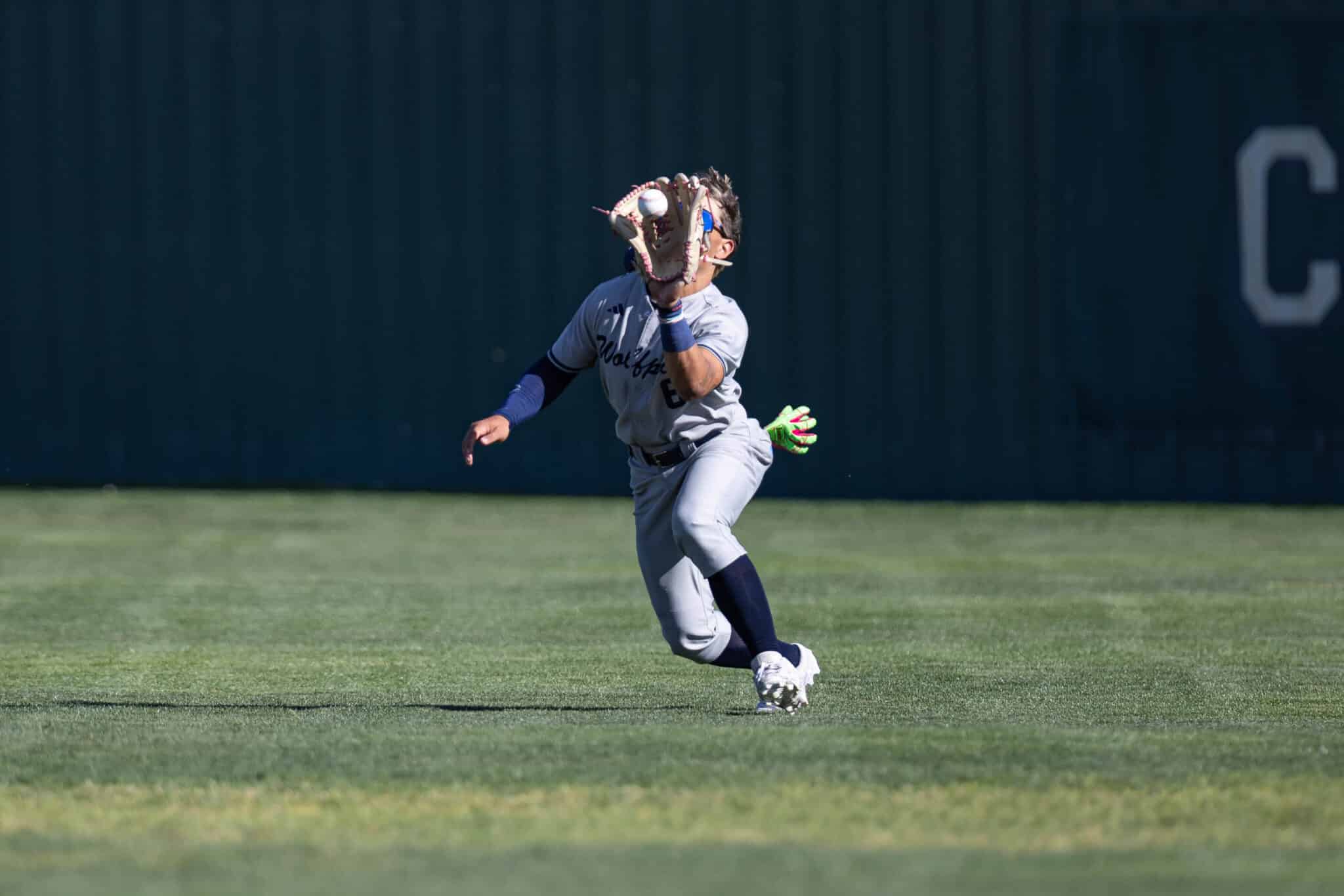 Baseball fielder in a gray uniform reaches with his gloved hand to catch a ball on a grassy field.