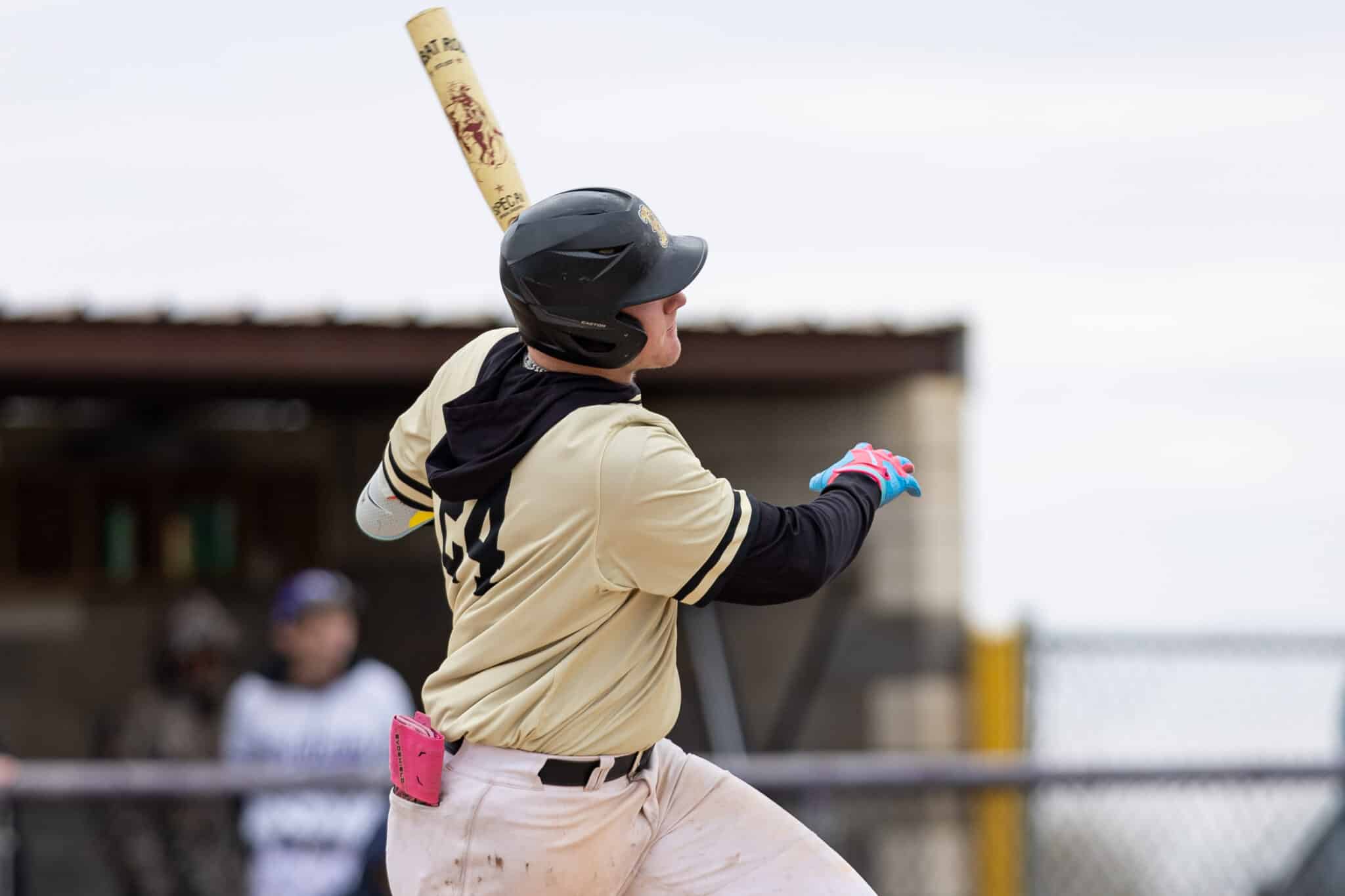 Baseball player in a beige uniform swings a wooden bat during a game, wearing a black helmet and blue-pink gloves.