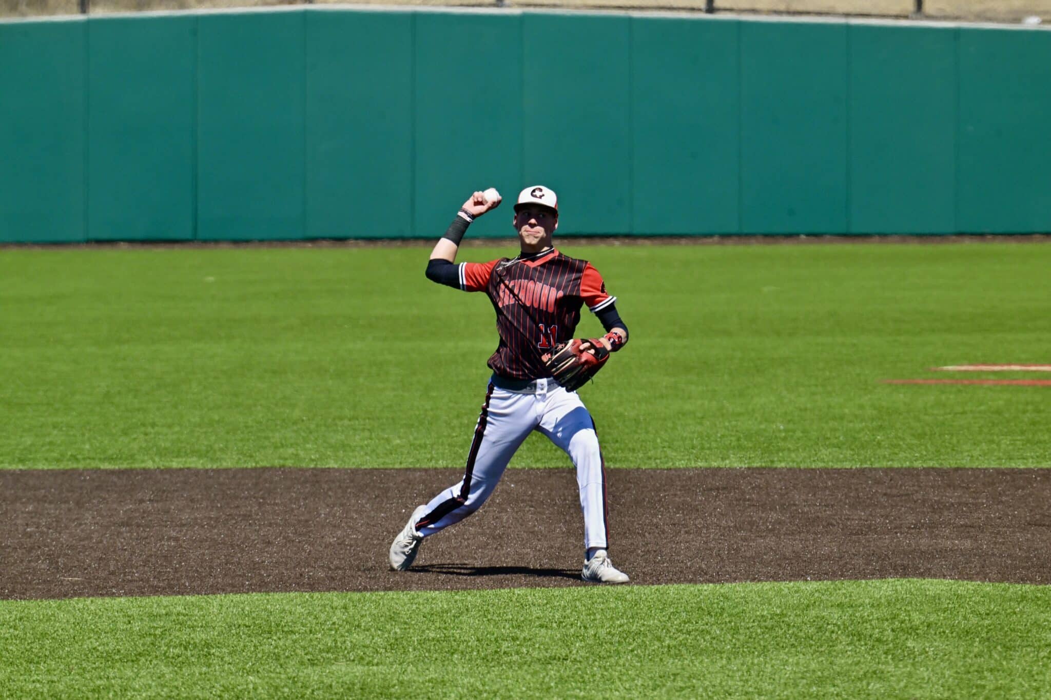 Baseball pitcher in red and black uniform delivering a pitch on the mound with a green outfield wall behind him.