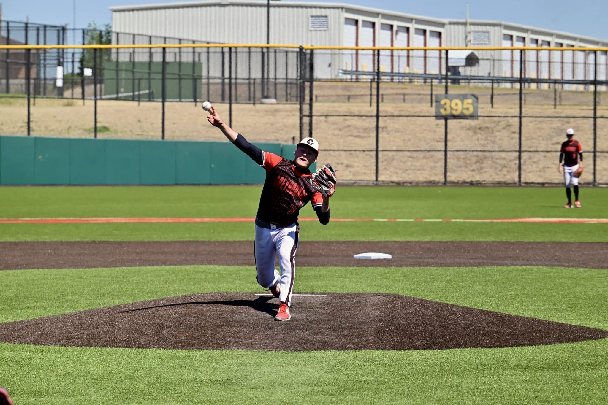 Baseball pitcher in red and black uniform throws from the mound on a sunny field.