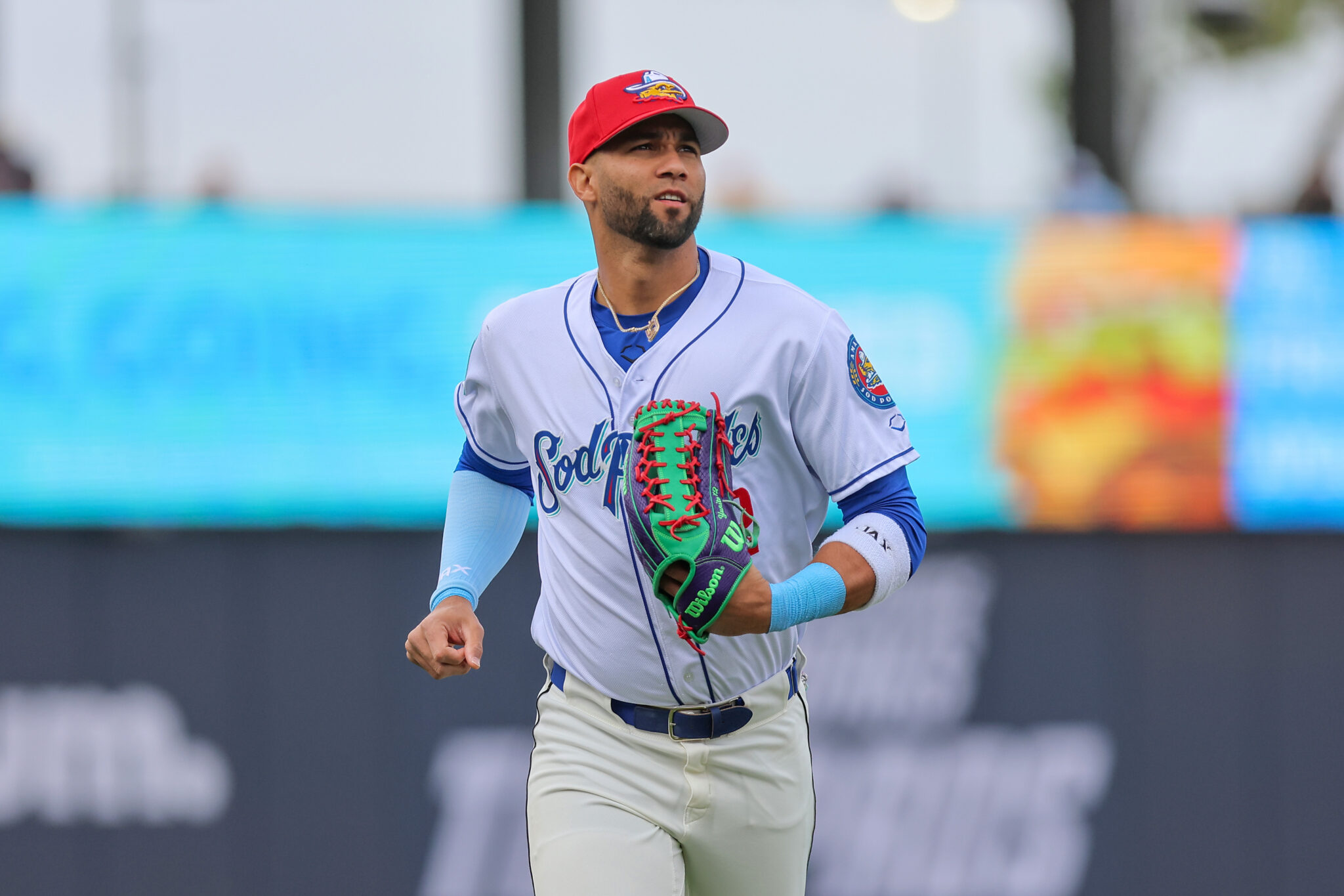 Baseball player in a white and blue uniform running with a green and red glove on his left hand, red cap on, blue sleeves visible in the foreground.