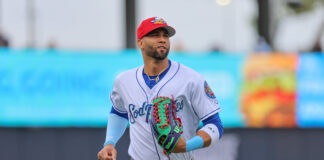 Baseball player in a white and blue uniform running with a green and red glove on his left hand, red cap on, blue sleeves visible in the foreground.
