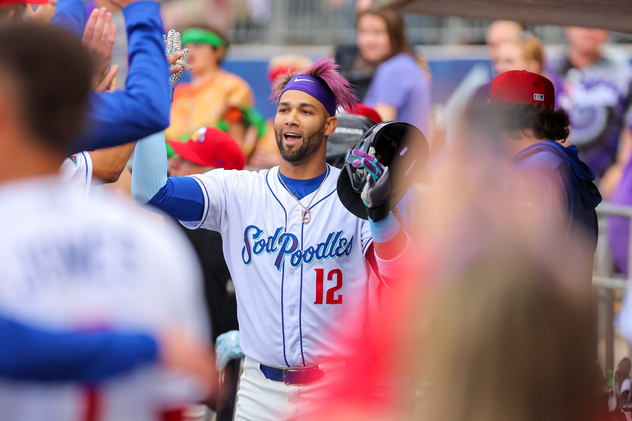 Baseball player with a purple headband and beard high-fives fans while holding a helmet, wearing a white and blue uniform number 12.