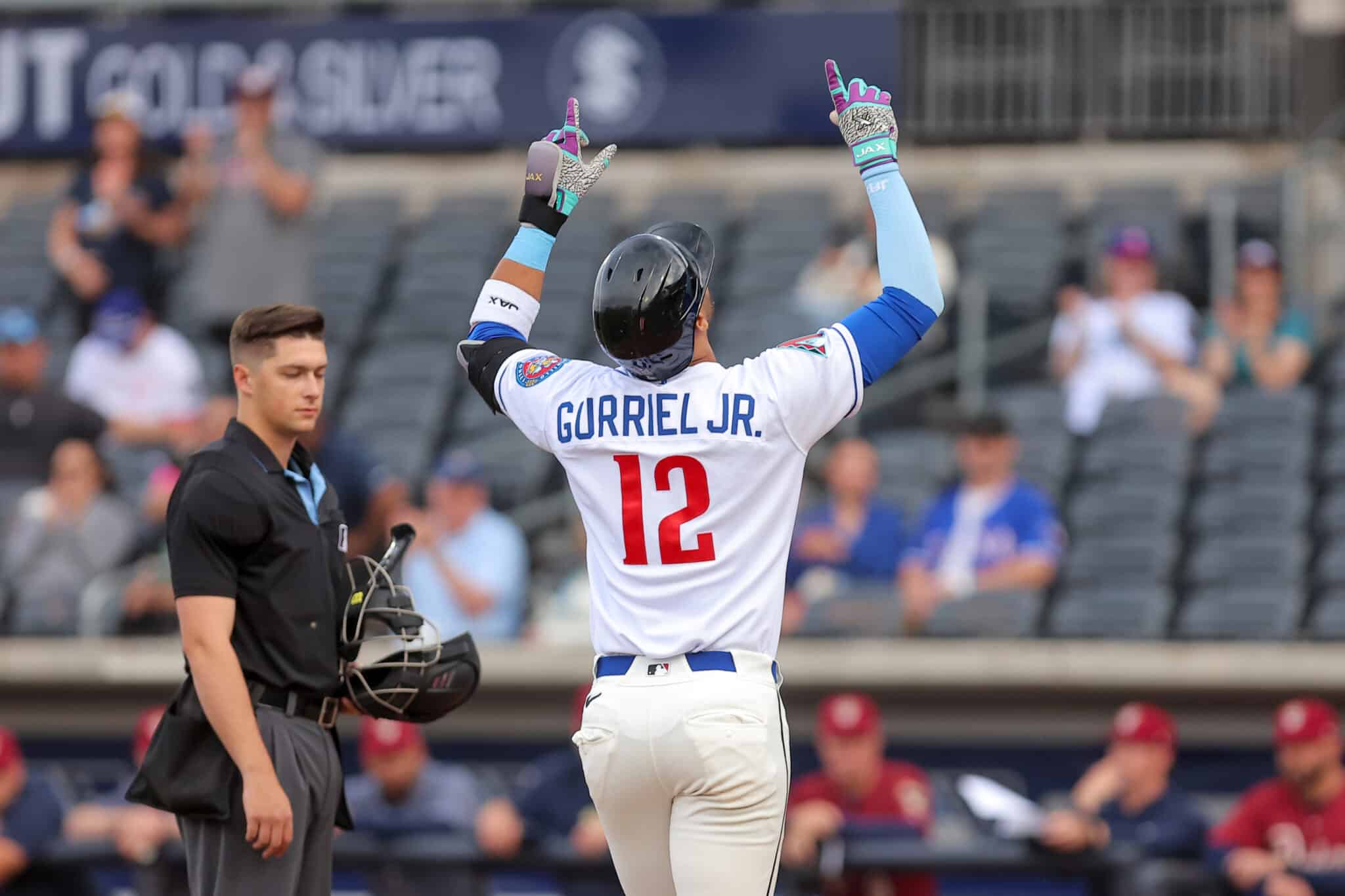 Baseball player Gurriel Jr., wearing white jersey number 12, raises both arms in celebration as the umpire stands nearby on the field.