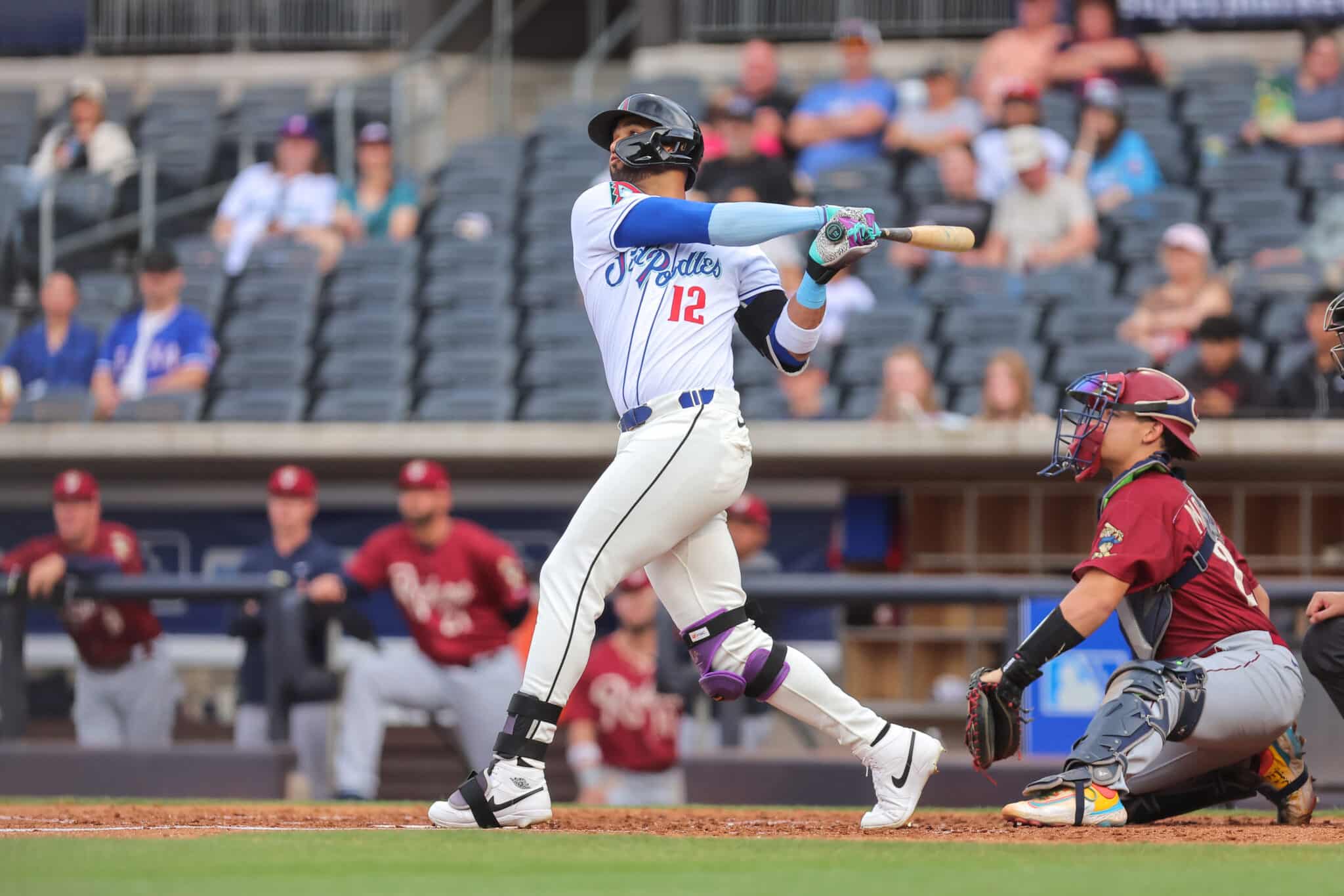 Baseball batter in white and blue swings at a pitch, with a catcher in maroon crouched behind him and fans in the stands behind.