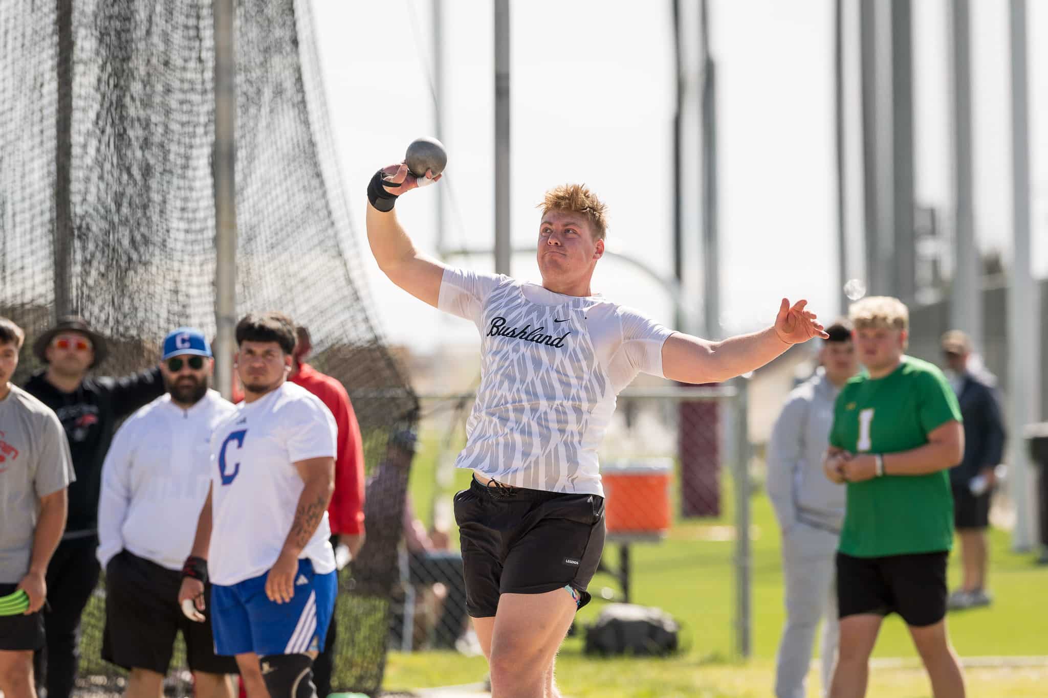 Male athlete in a white shirt (Bushland) releasing a shot put during a field event, with spectators behind a safety net.