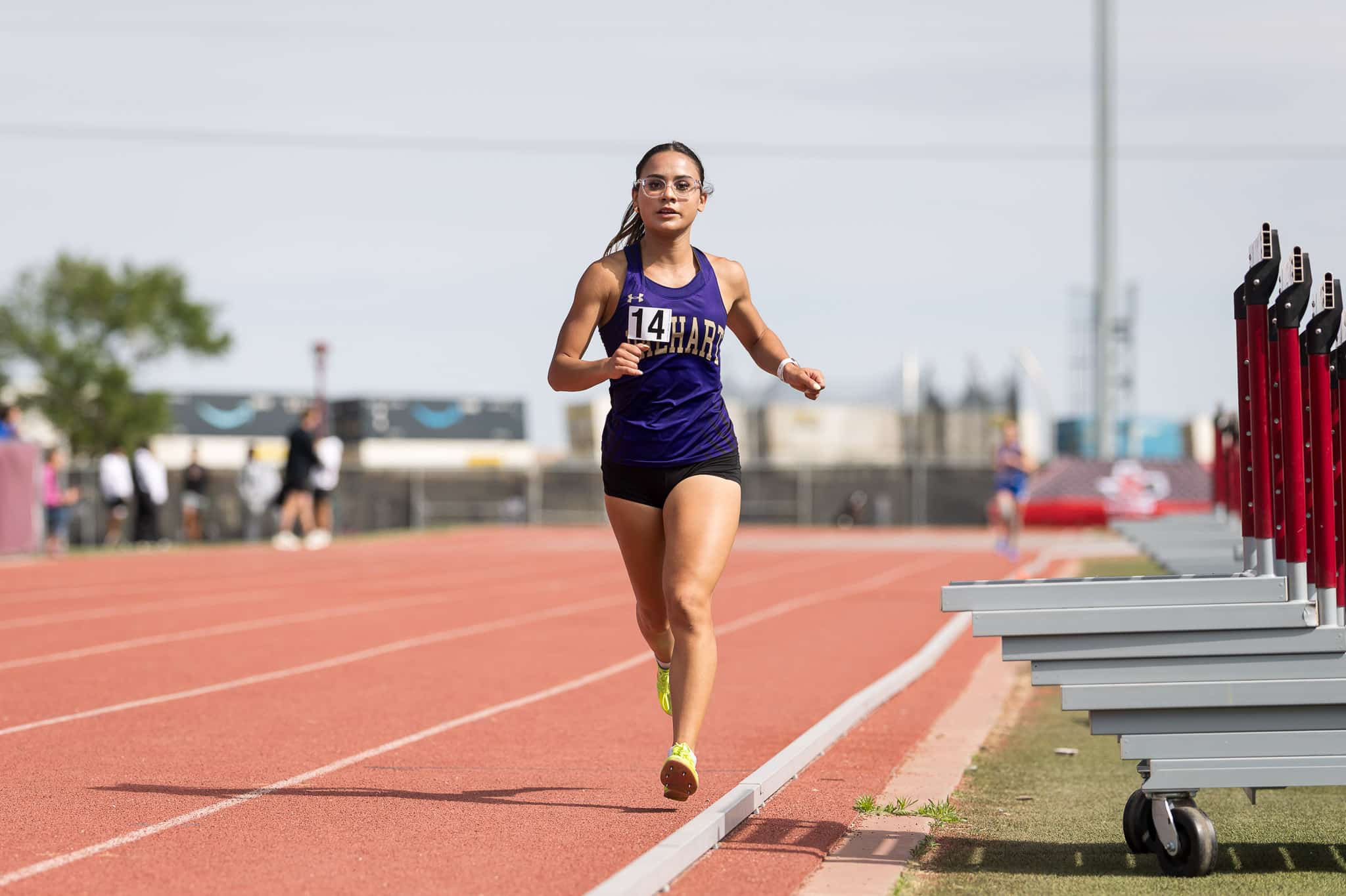 Female athlete in a purple top and black shorts sprinting on a red track wearing bib 14.
