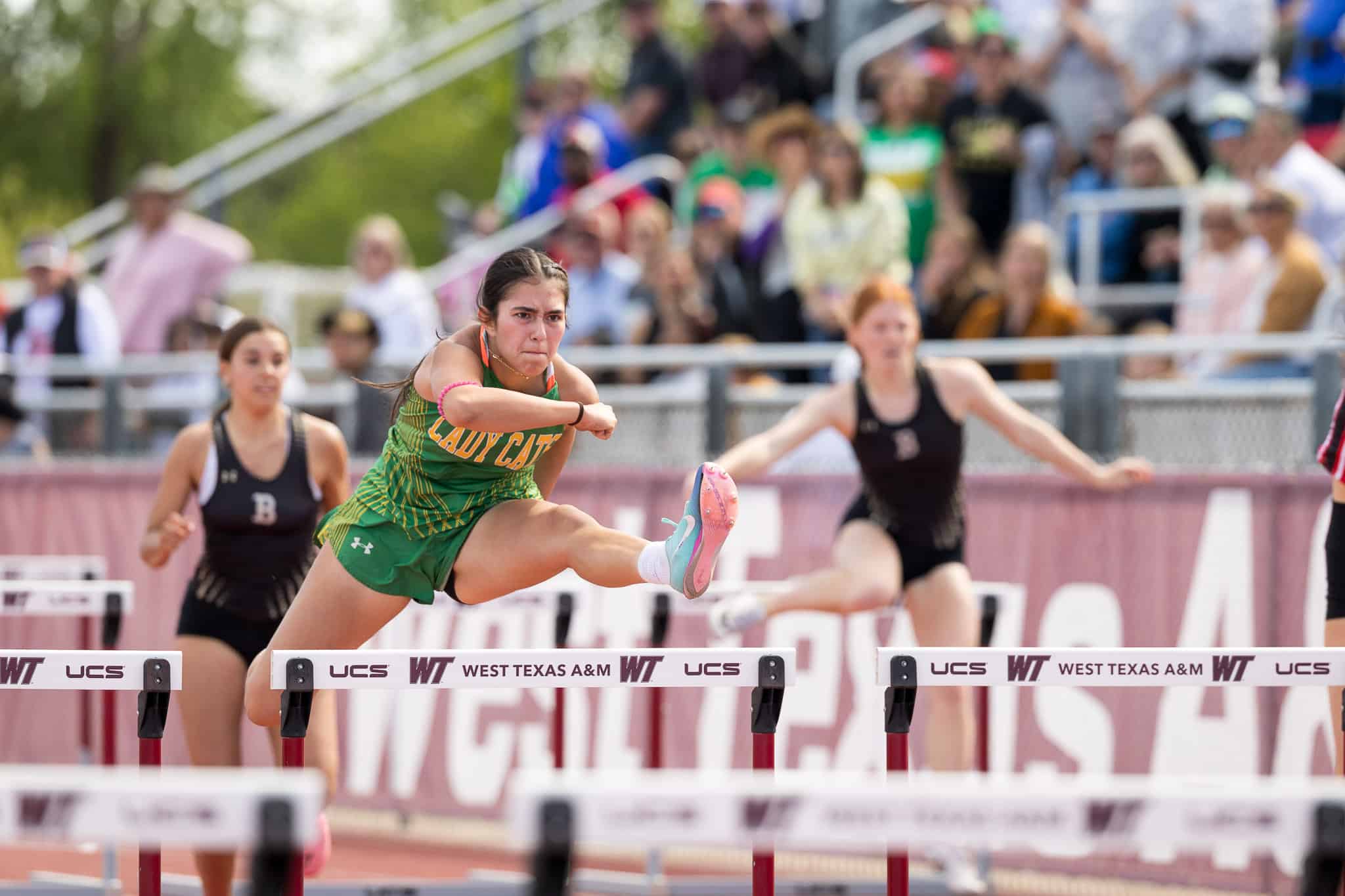 Female athlete in a green uniform clearing a hurdle mid-race at a track meet, with blurred crowd in the stands behind her.