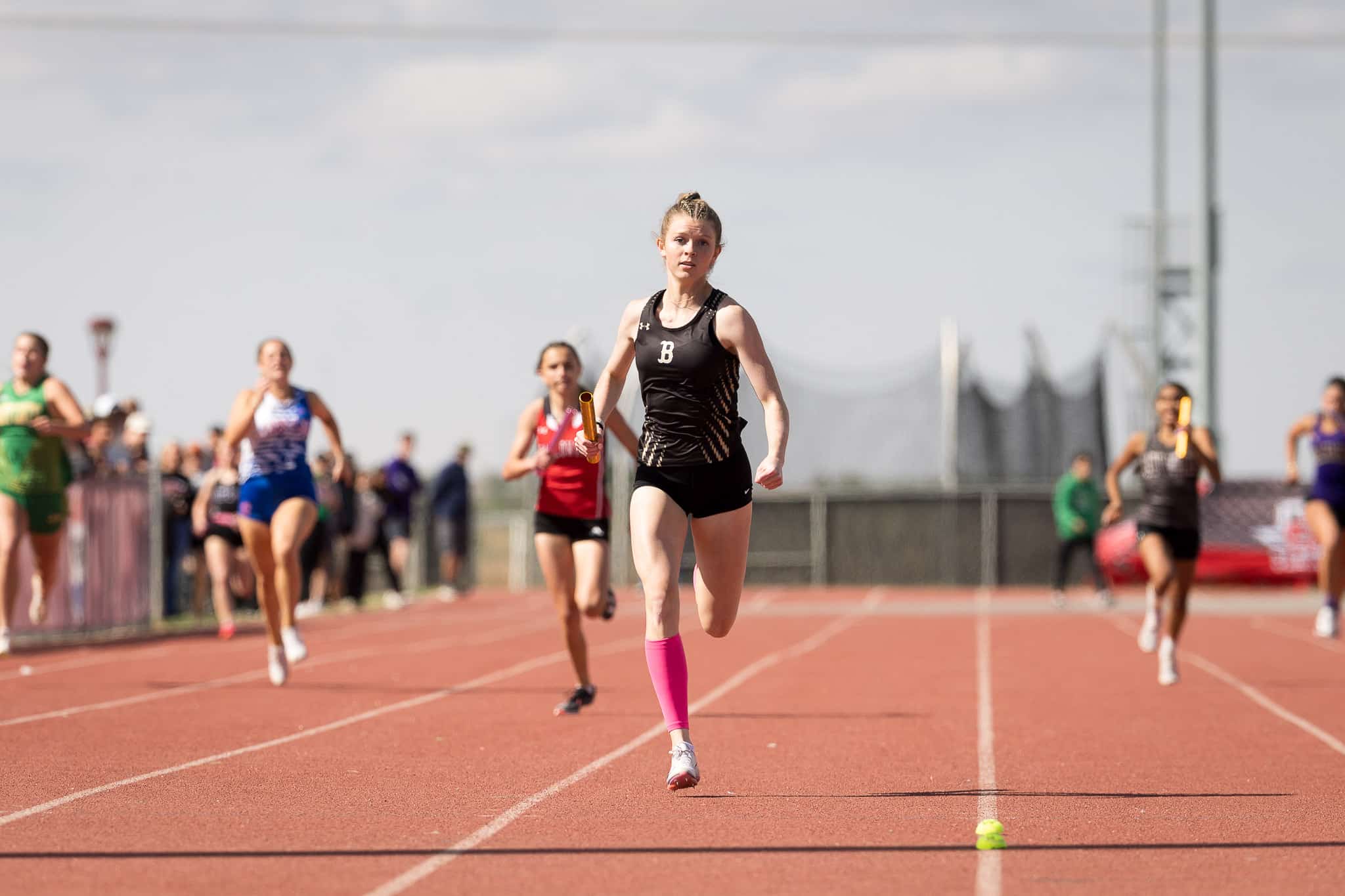 Female sprinter in a black uniform leads a relay race on a red track, holding a baton as other runners follow behind.