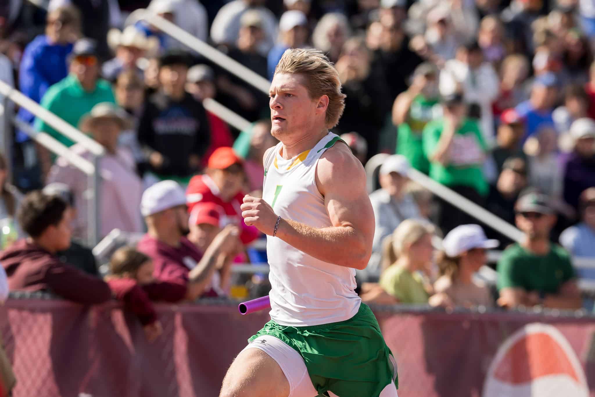 Male runner in white and green, sprinting with a purple relay baton in a crowded race, spectators behind barriers.